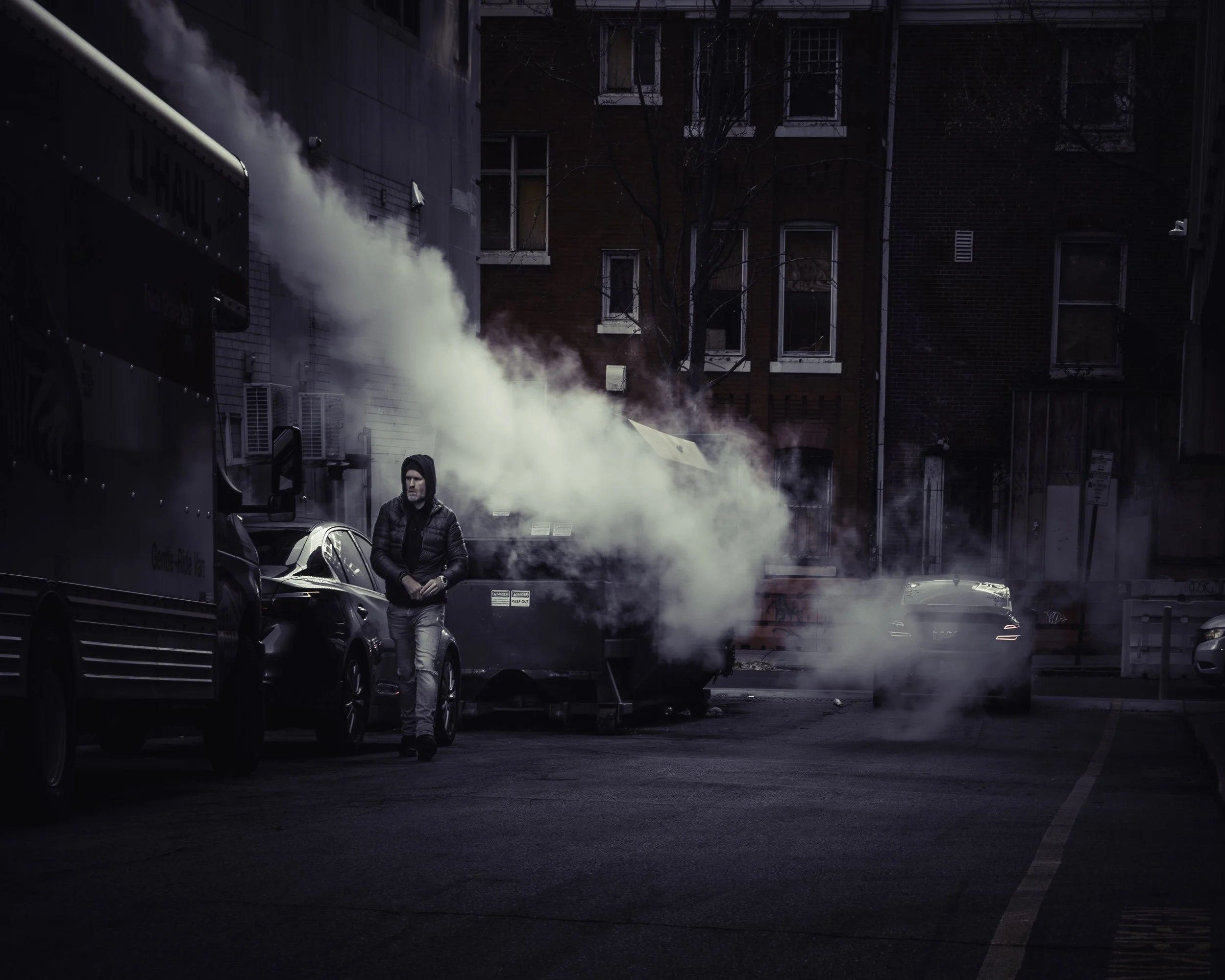 Steam Alley
Hooded man walking through steam in a dark alley beside parked cars and a U-Haul truck.