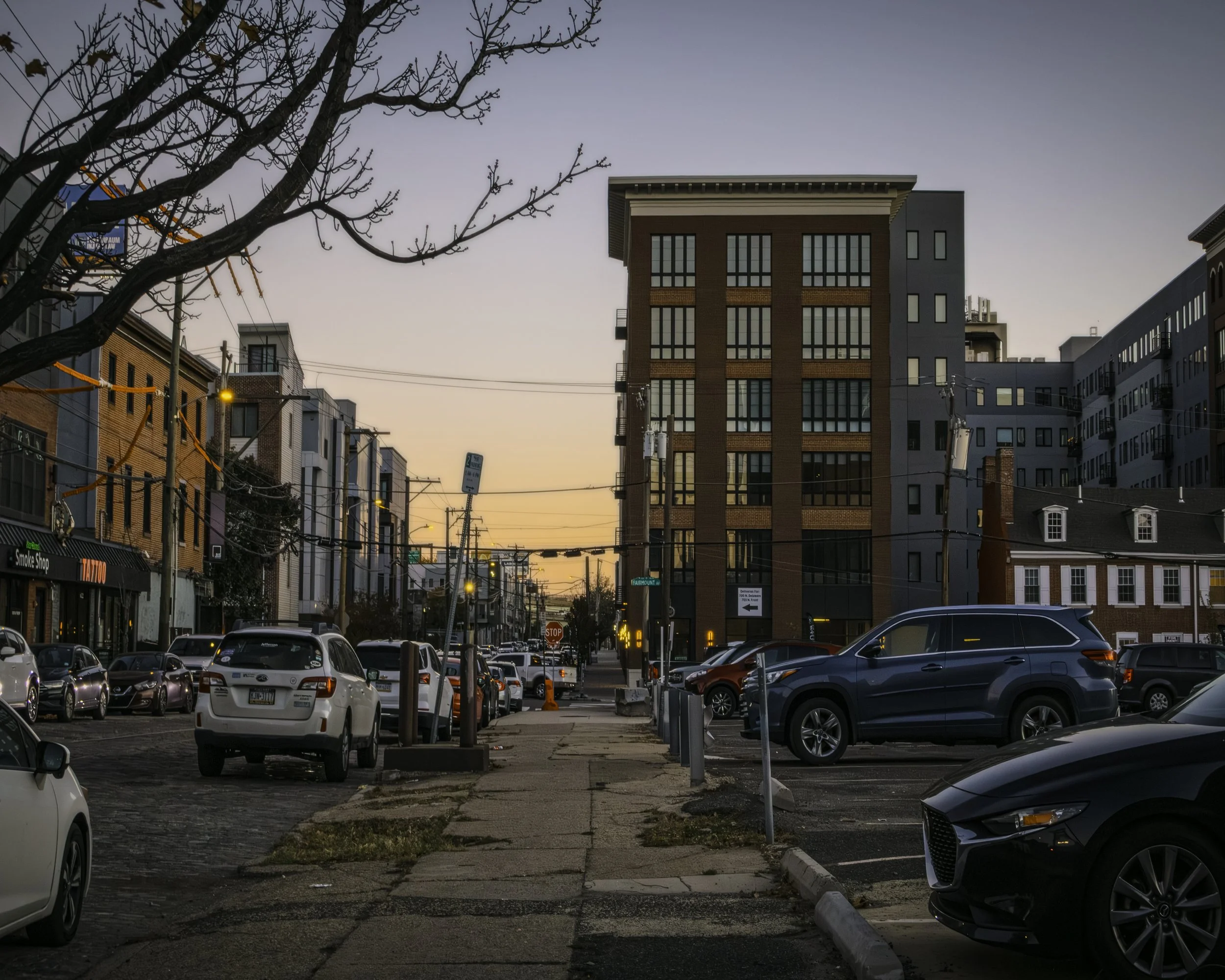 Street scene in Philadelphia’s Northern Liberties at dawn, with parked cars, a tall brick apartment building, and a soft orange-blue sky creating a calm urban atmosphere.