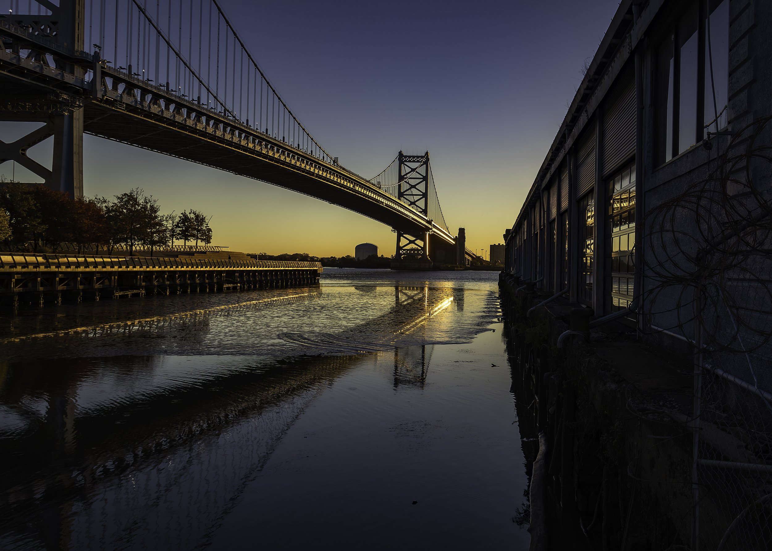 River Gold
Wide sunrise view of the Ben Franklin Bridge spanning calm river water, golden light reflecting off the bridge, autumn trees, and warehouse windows, creating a sense of expansive stillness.