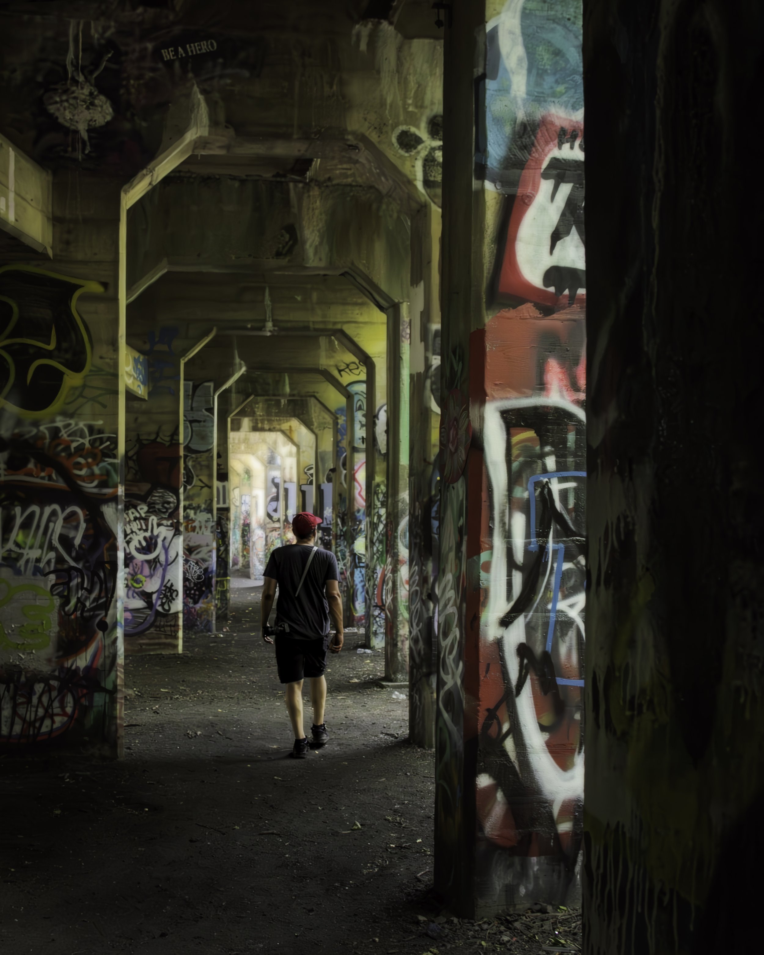 Through the Ruins
A lone figure walks away from camera through a long corridor of massive graffiti-covered concrete columns in an abandoned Philadelphia waterfront pier structure, receding arches glowing green in the distance, industrial decay framin