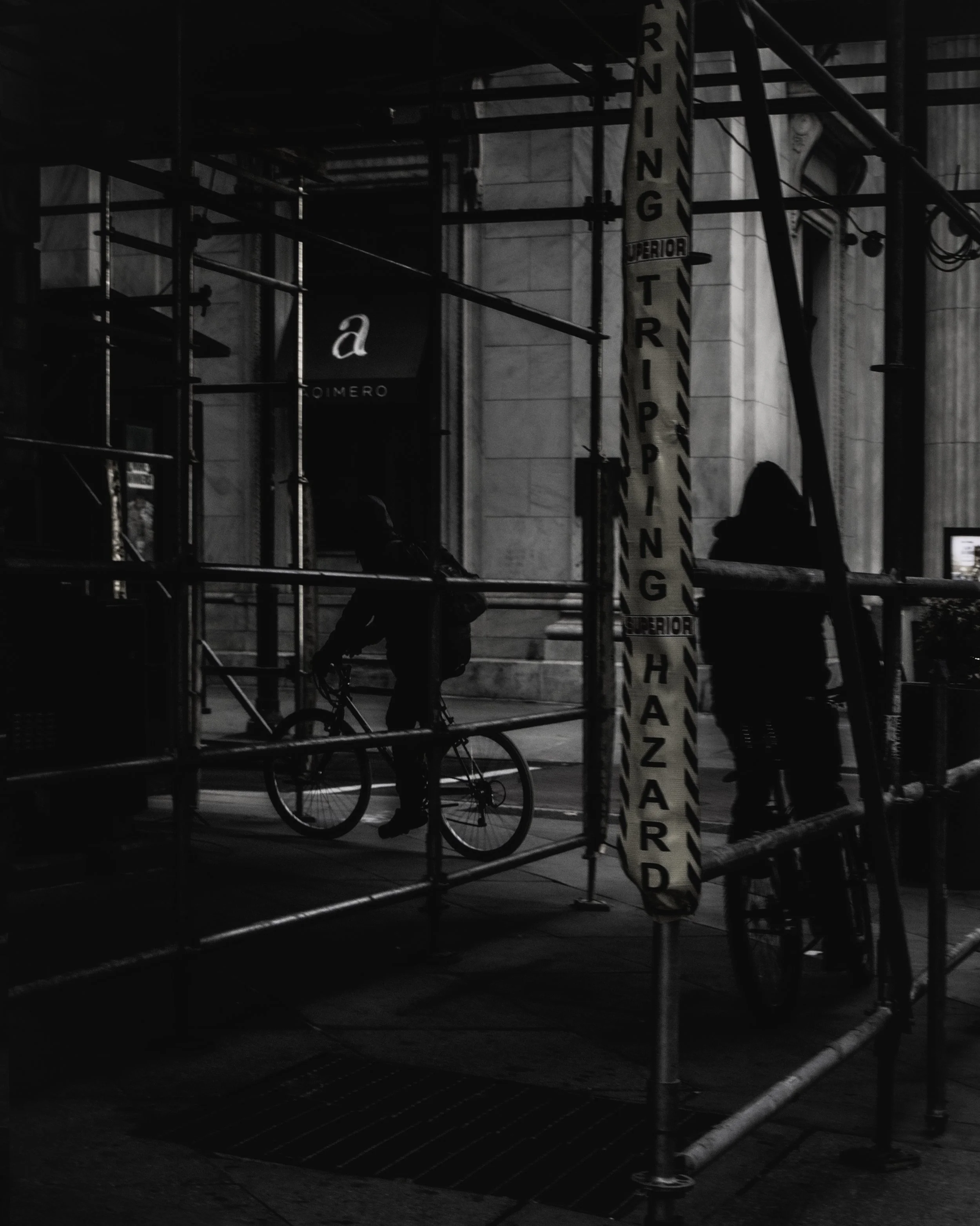 Trip Hazard
Dark street scene in downtown Philadelphia with two cyclists framed by scaffolding and a vertical warning sign reading trip hazard.