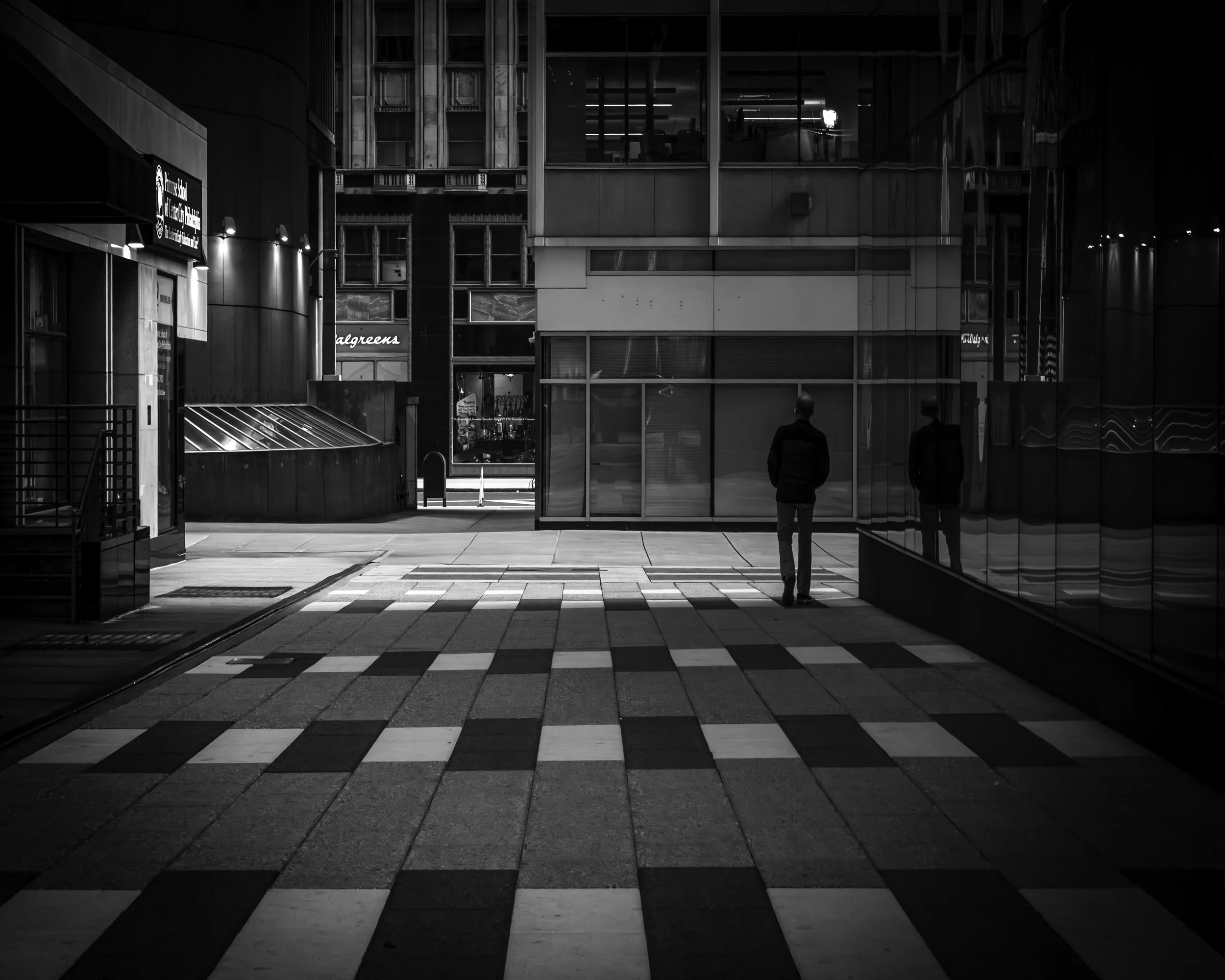 Glass and Stone
Black-and-white photograph of a lone person standing in a modern city passageway, with checkered paving, glass reflections, and office windows glowing above.