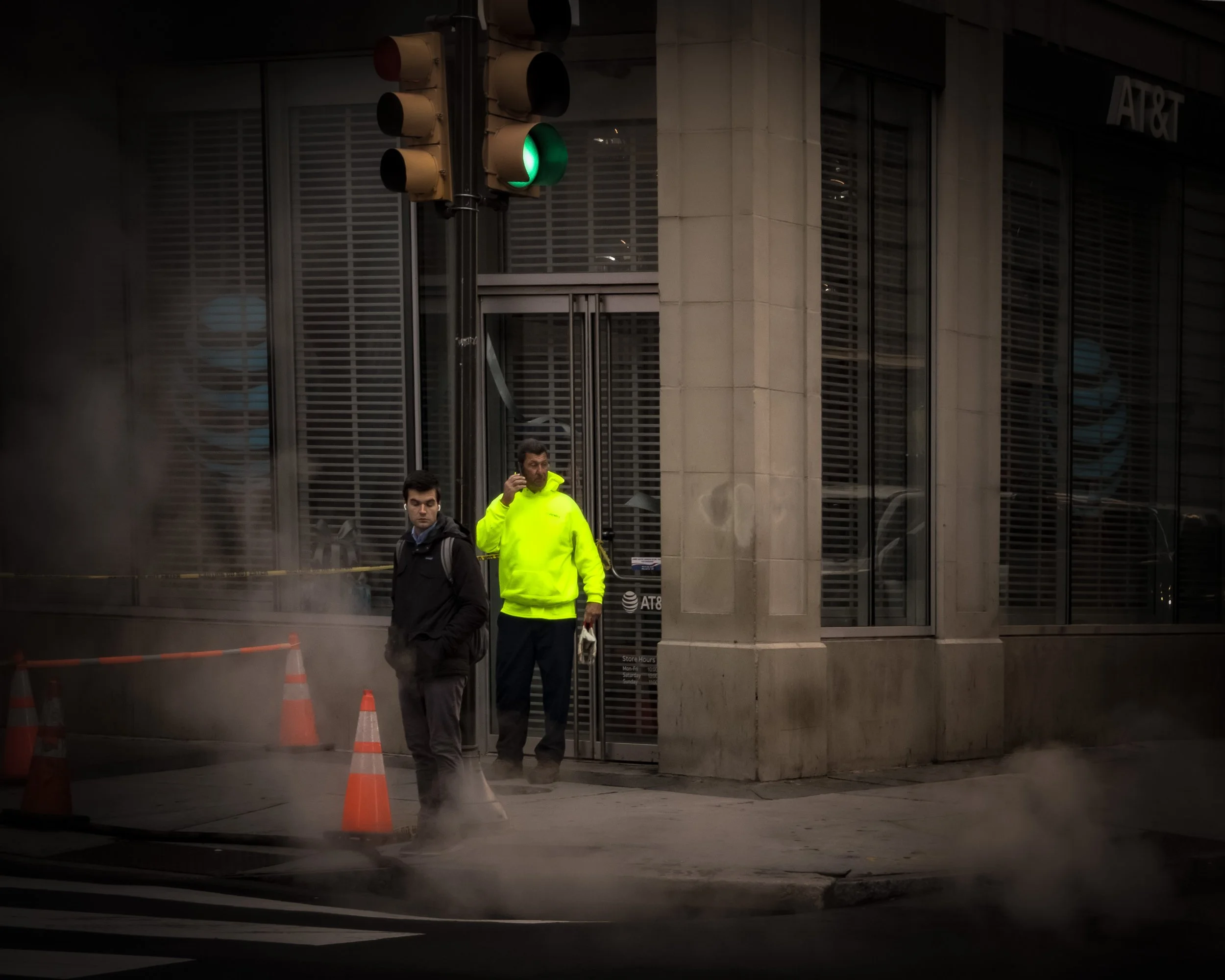 Corner in Haze
Man in a bright neon jacket stands in a doorway beneath a green traffic light while steam rises around orange cones at a street corner.
