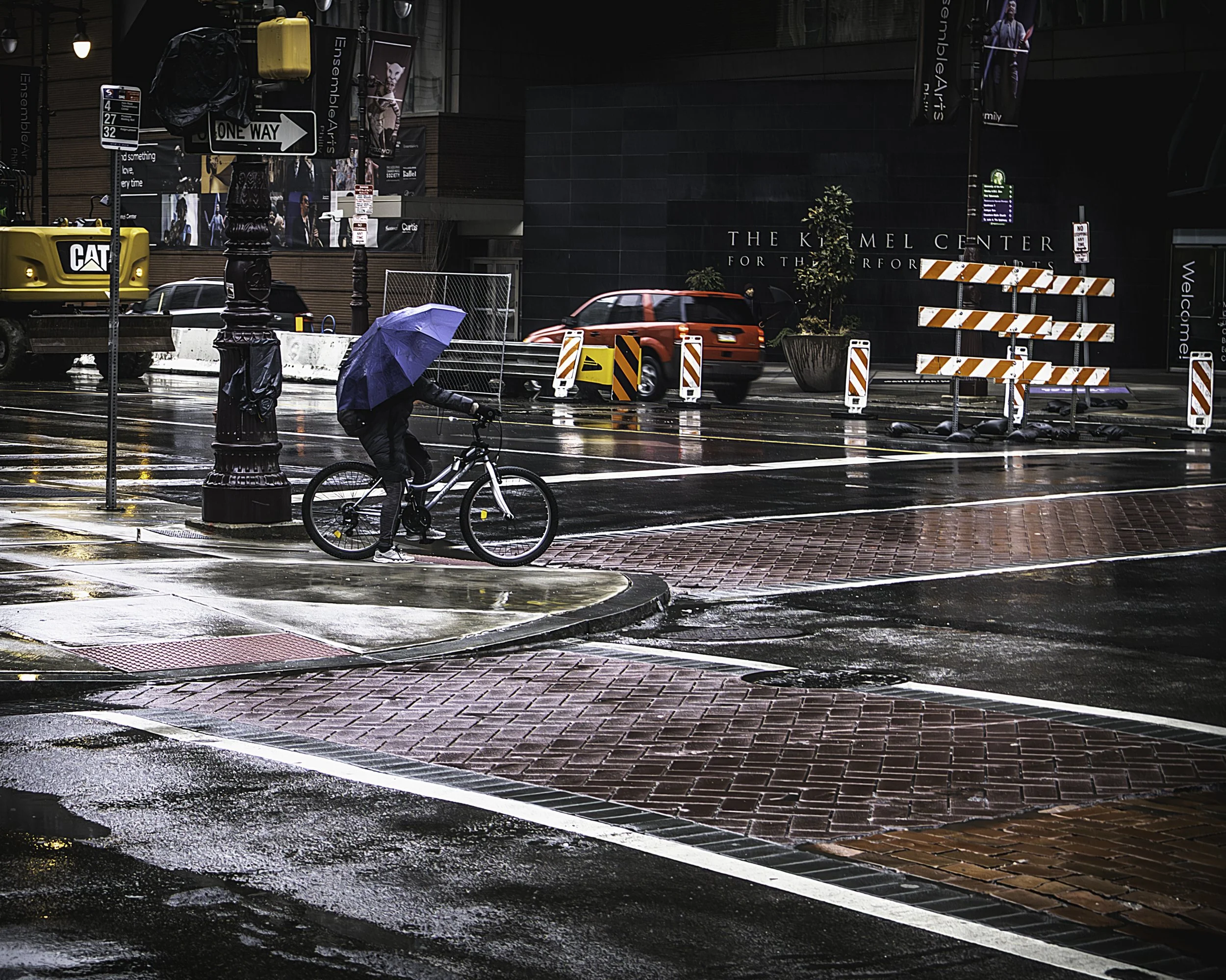 Waiting at the Kimmel
Cyclist pauses at a rain-slick corner beneath a purple umbrella across from the Kimmel Center for the Performing Arts.