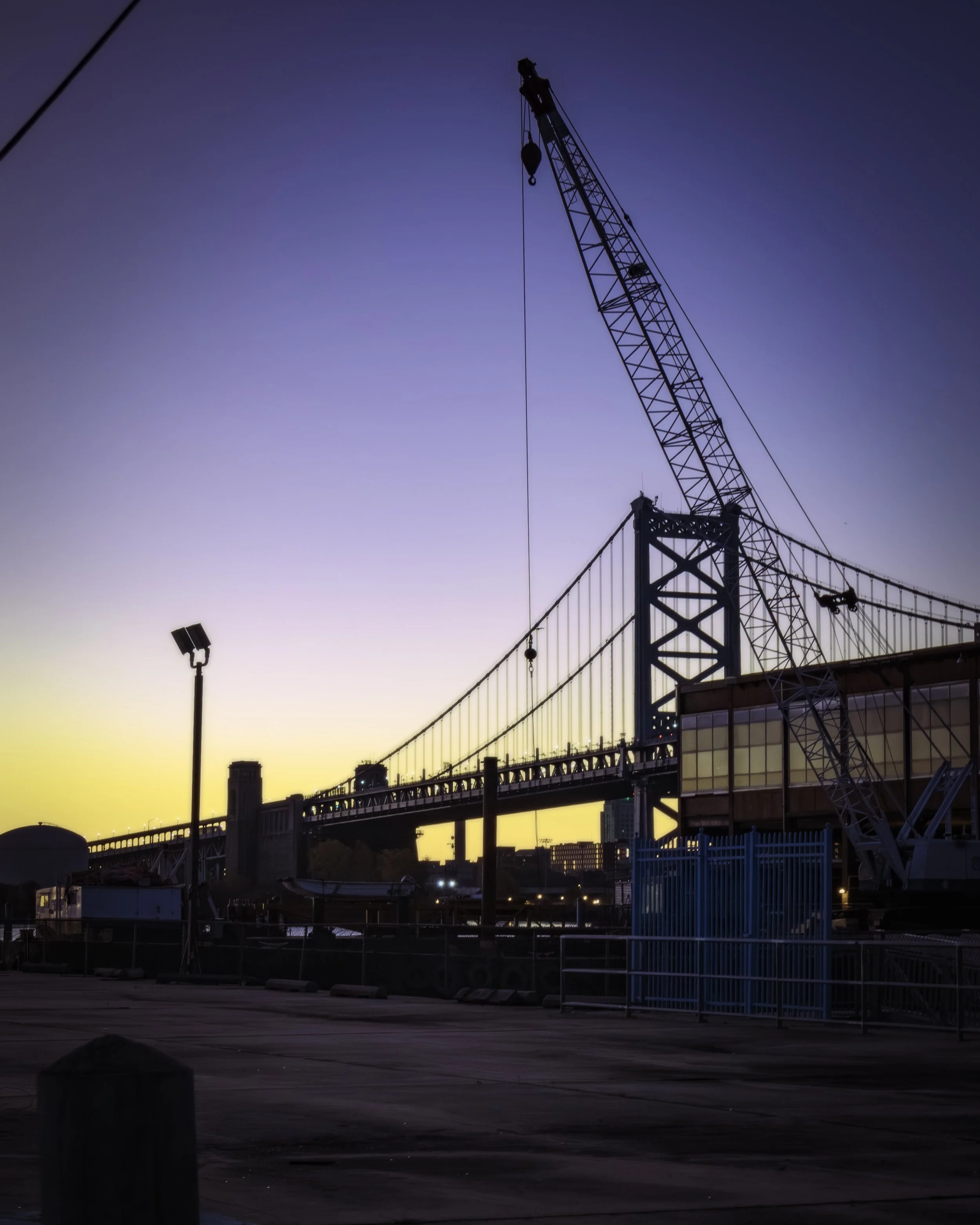 Still and Calm Begins the Day
Crane rises beside the Benjamin Franklin Bridge at dawn, with a pale sky above an open waterfront work area.