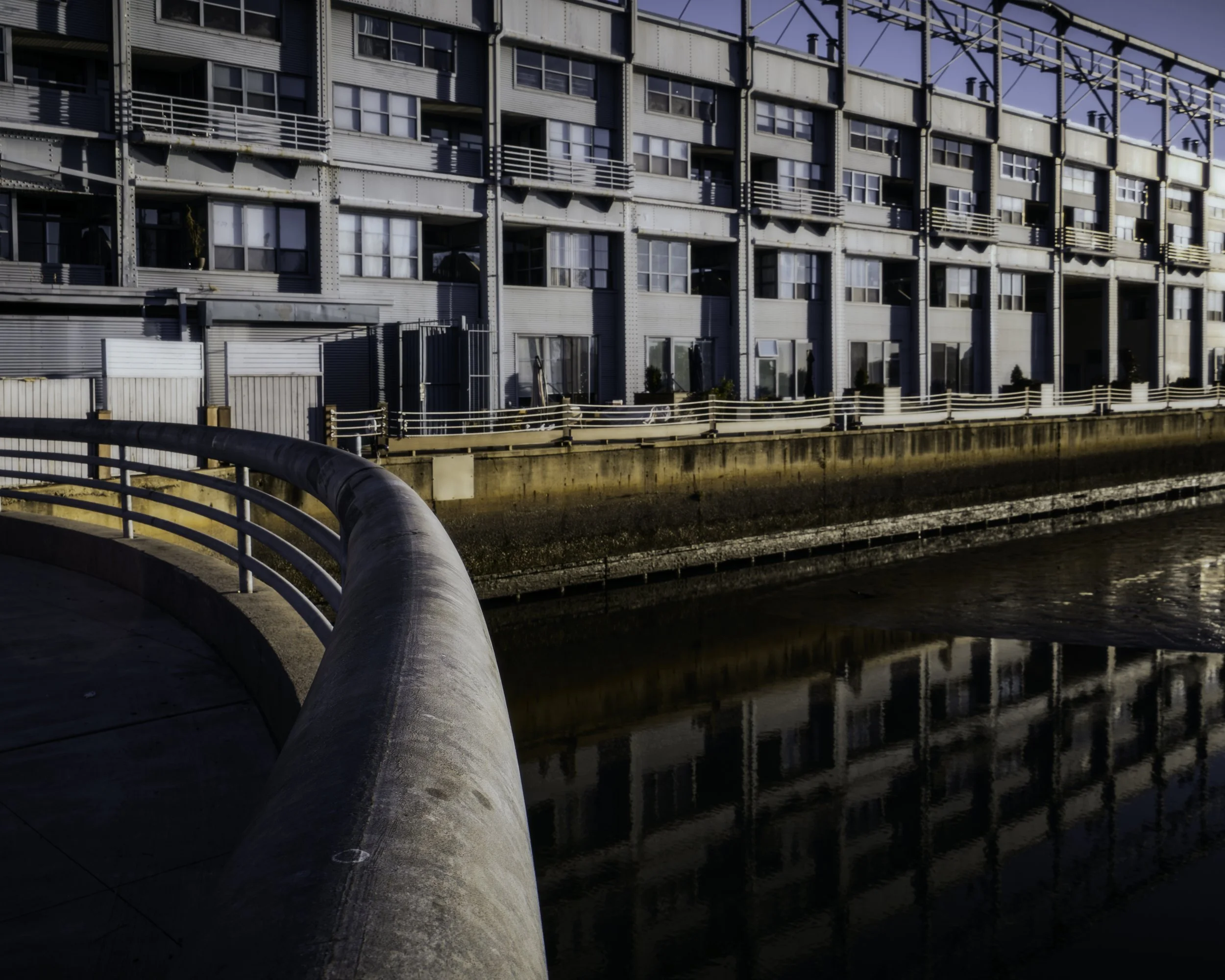 Lines and Reflections
Curving waterfront railing leads toward a long residential building, with reflections mirrored in the narrow channel below.