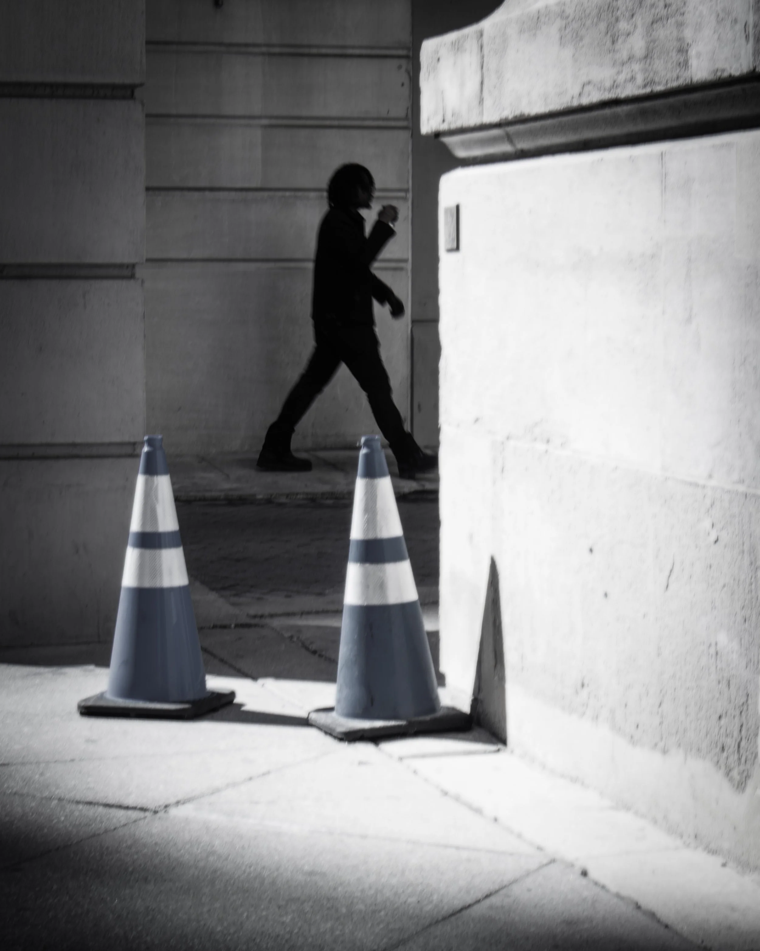 Passing the Cones
Black and white photograph of a silhouetted pedestrian walking behind two blue traffic cones beside a bright stone wall at The Bellevue parking garage.