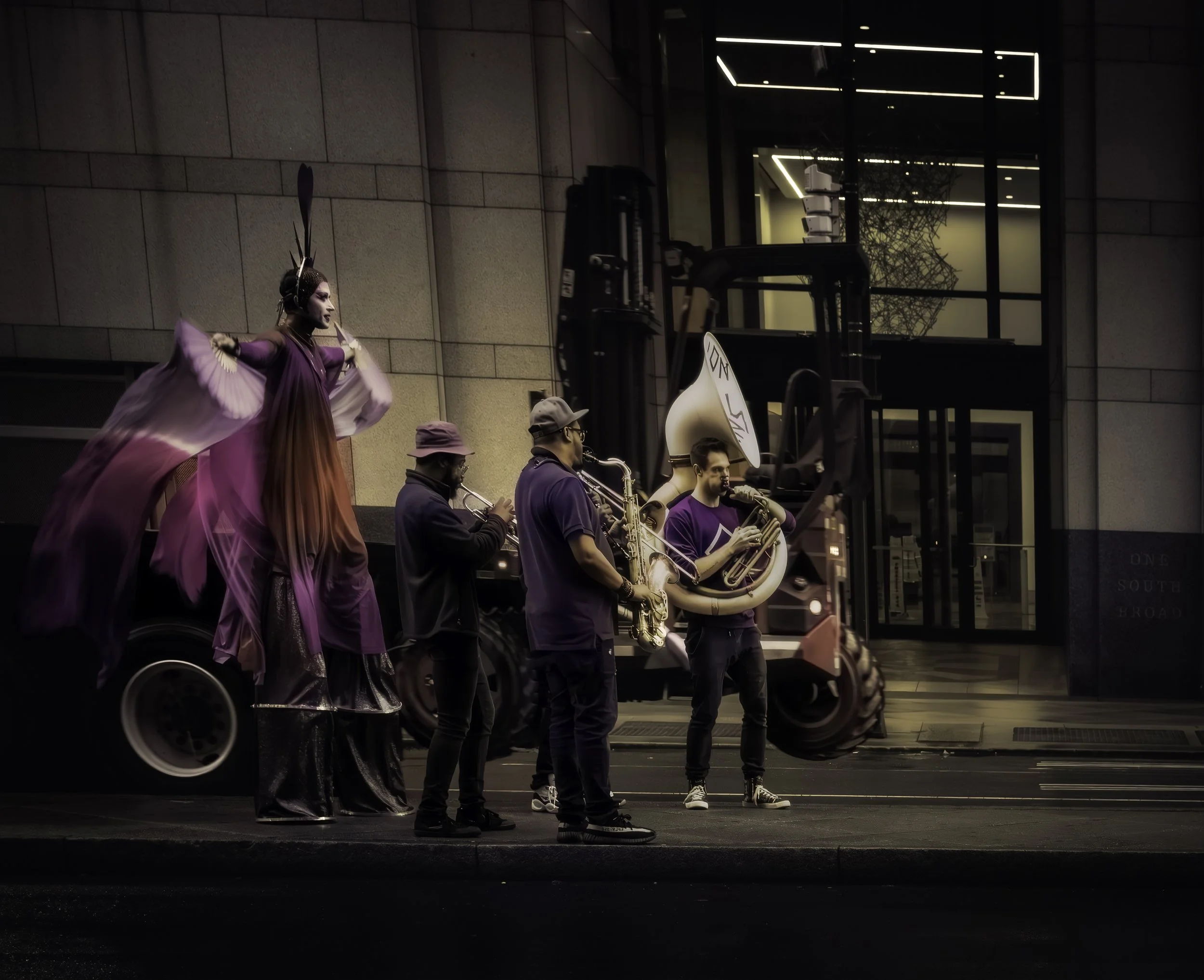 Midnight Brass and Velvet Wings
Stilt walker in a flowing purple costume performs beside a small brass band on a city street.