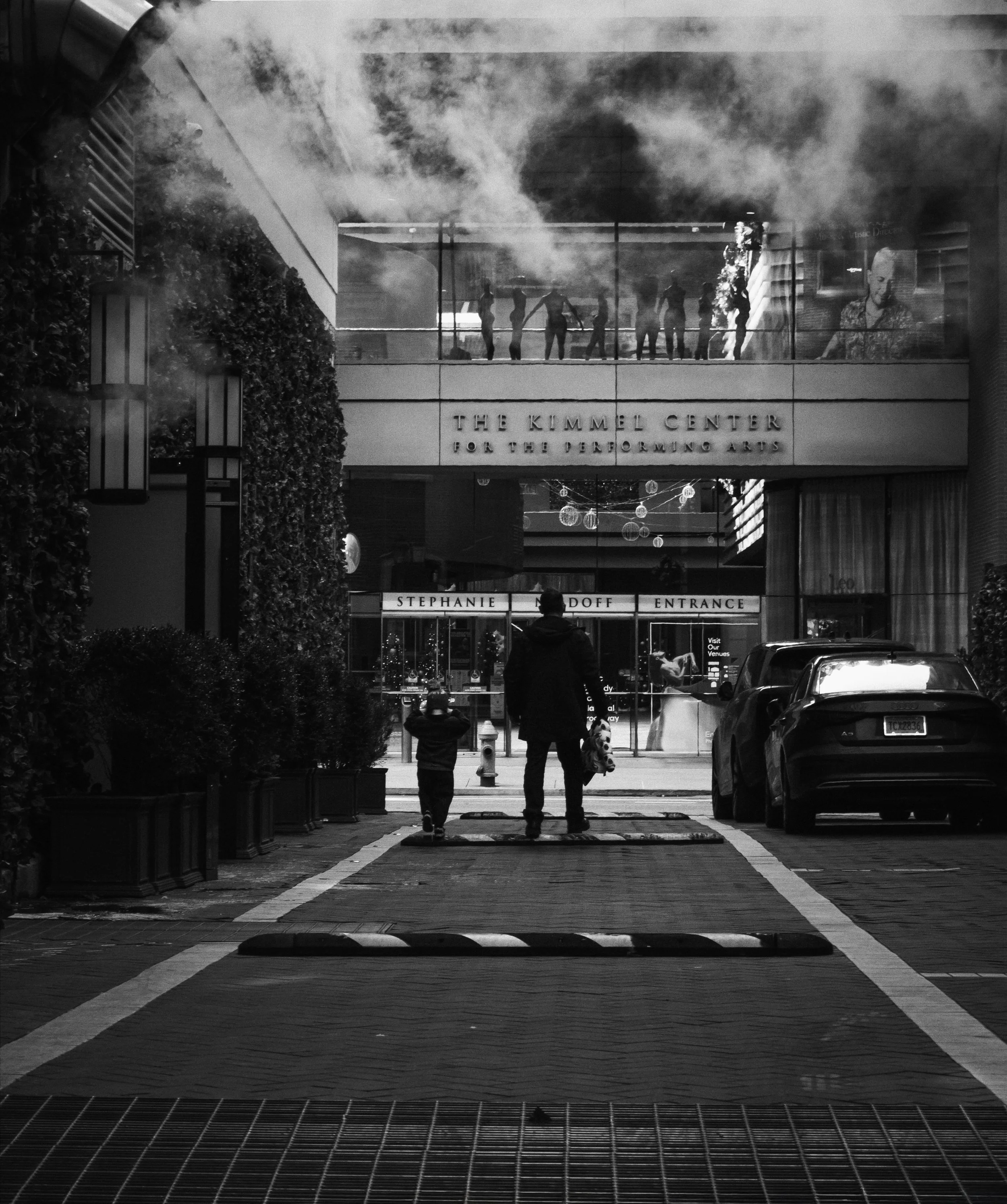 The Alley to Broad Street
Adult and child standing in an alley facing a glass-fronted building, with steam overhead and silhouetted figures visible in upper windows.