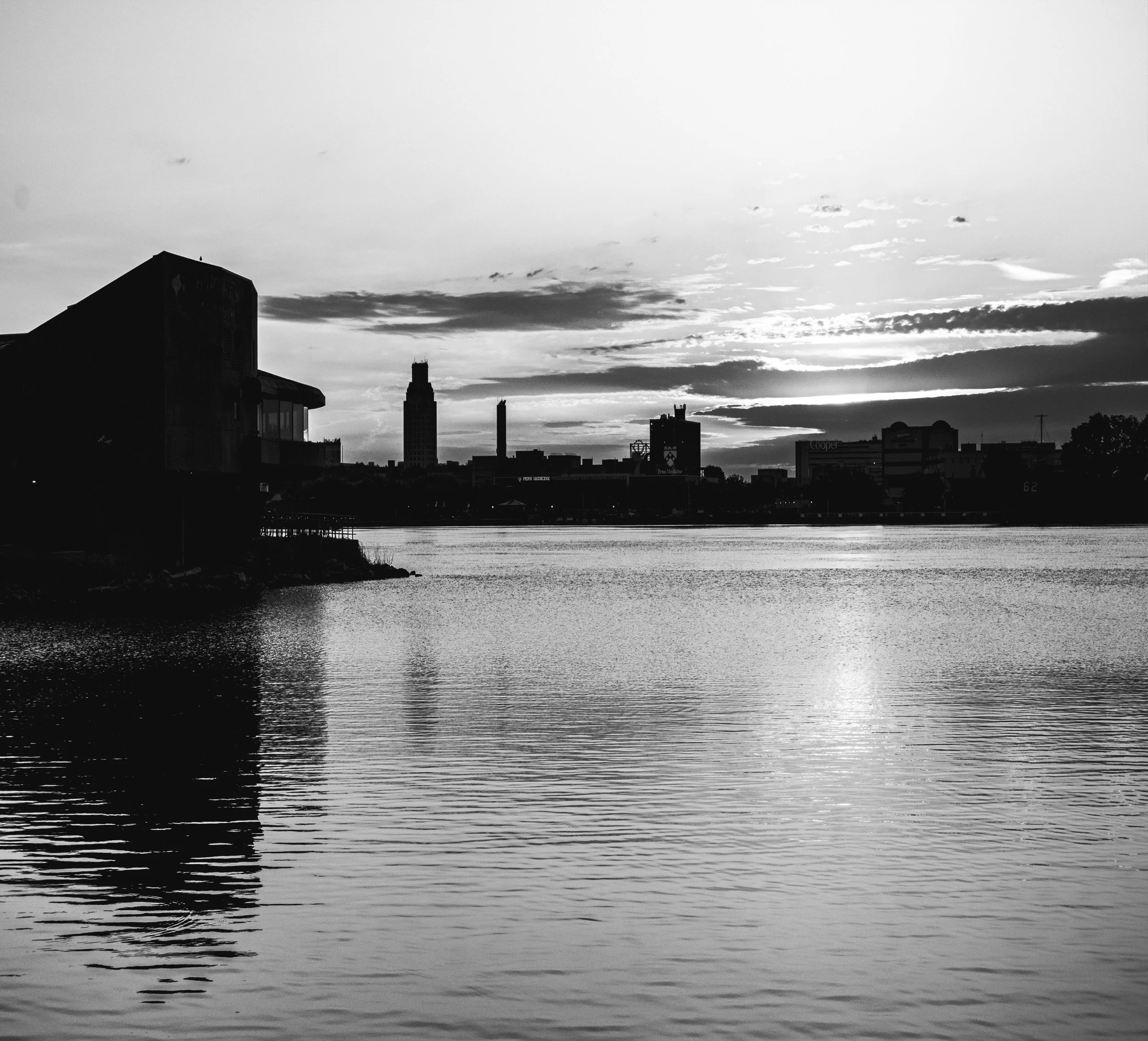 Black and white cityscape silhouette across water at dawn, dramatic cloud formations in sky, buildings reflected in rippling water surface, urban waterfront scene