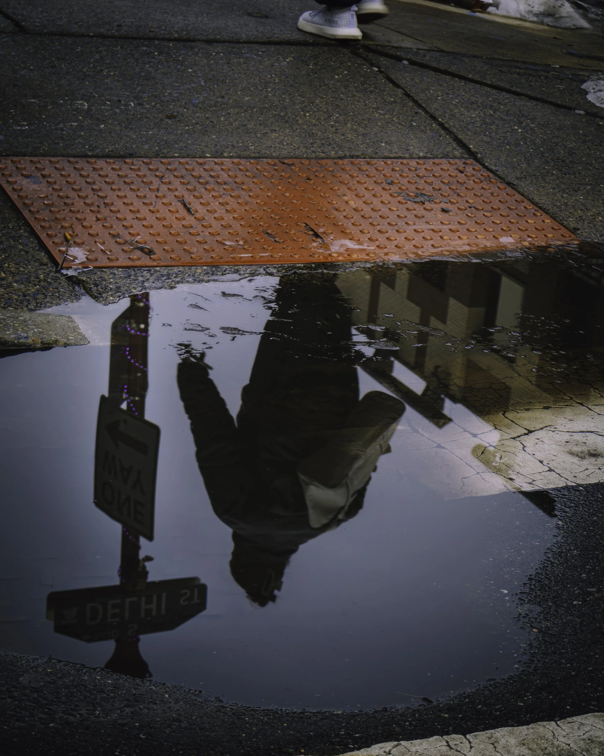 One Way in the Water
Puddle reflection of a pedestrian crossing near a tactile curb ramp, with a one-way sign and street corner mirrored in the water.