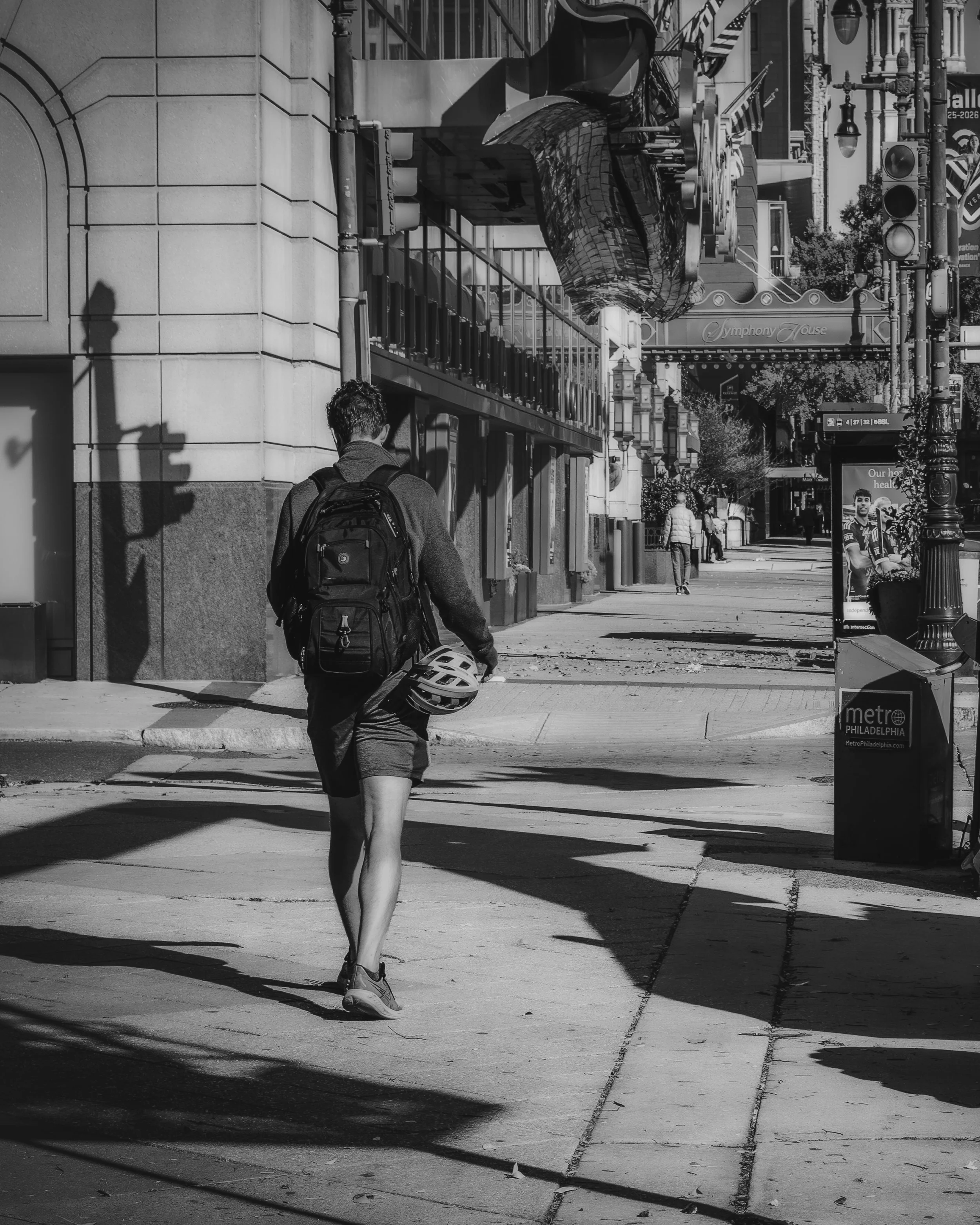 Backpack, Helmet, and a City of Shadows
Black and white street photograph on Philadelphia’s Avenue of the Arts, showing a pedestrian with a backpack and bicycle helmet walking beneath deep building shadows