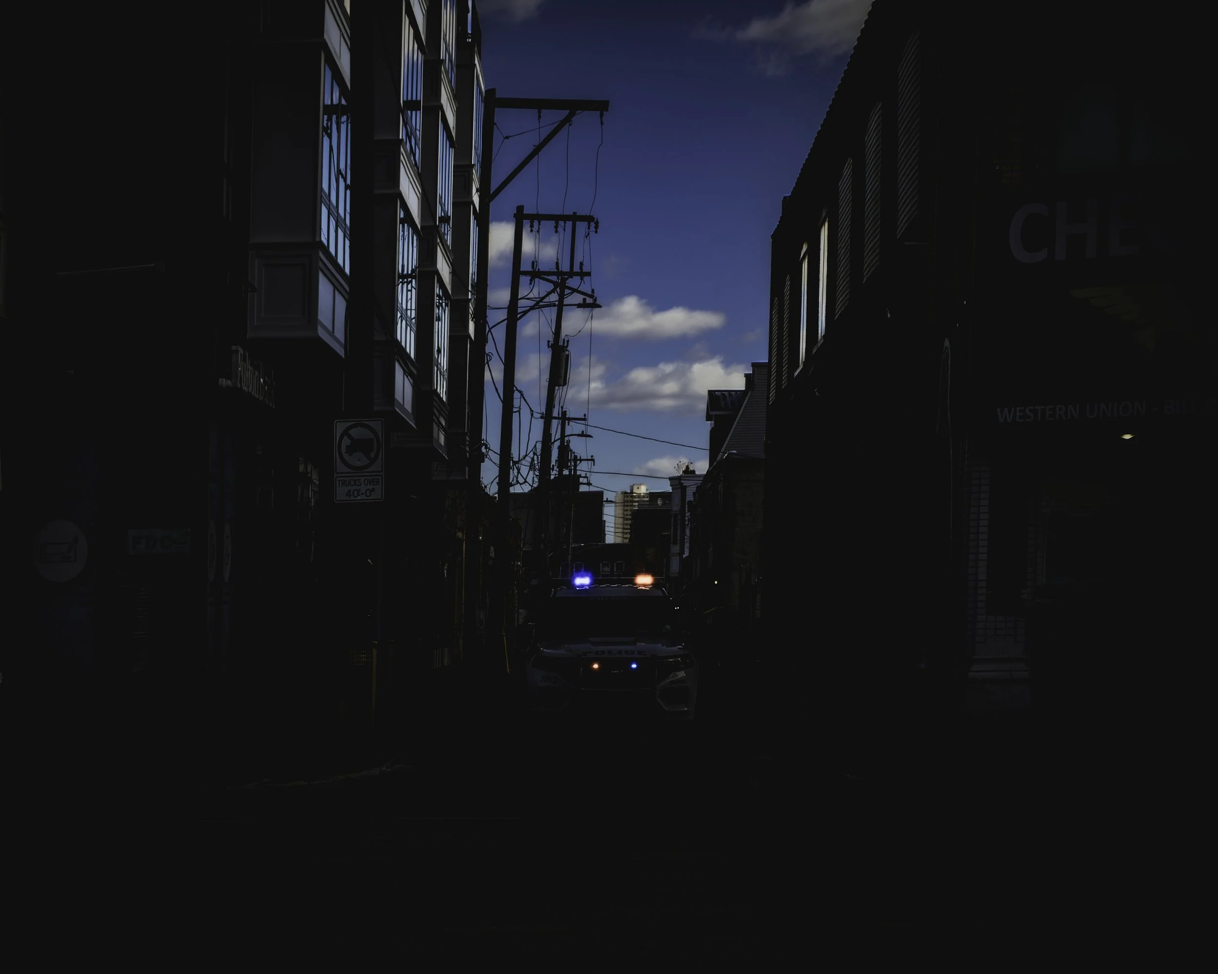 Flashing Blue Distance
Dark alley framed by buildings and utility poles, with a police vehicle showing flashing lights in the distance.