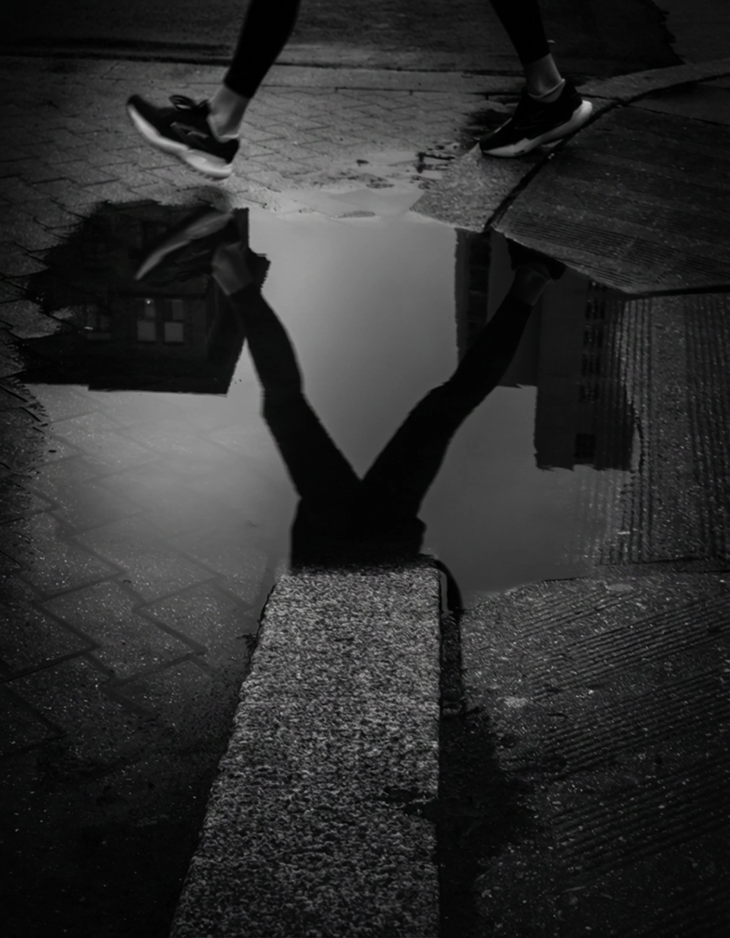 Crossing the Divide
Black and white street photograph of a pedestrian crossing beside a rain puddle, with the figure reflected in the water between textured curb cuts and sidewalk edges.