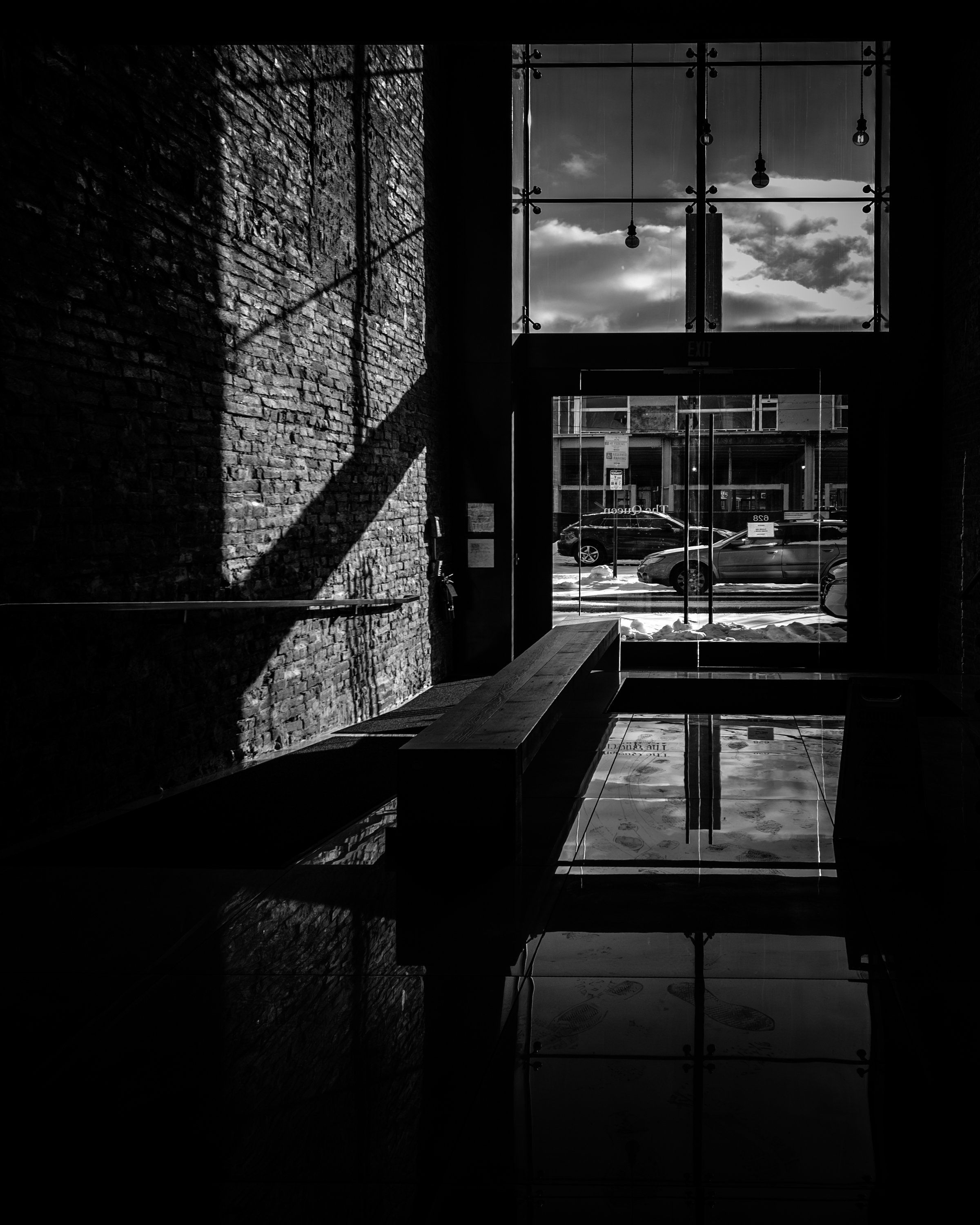 Winter Through Glass
Black and white photograph of a modern lobby with a sunlit brick wall, glass entrance doors, and reflections on a polished floor.