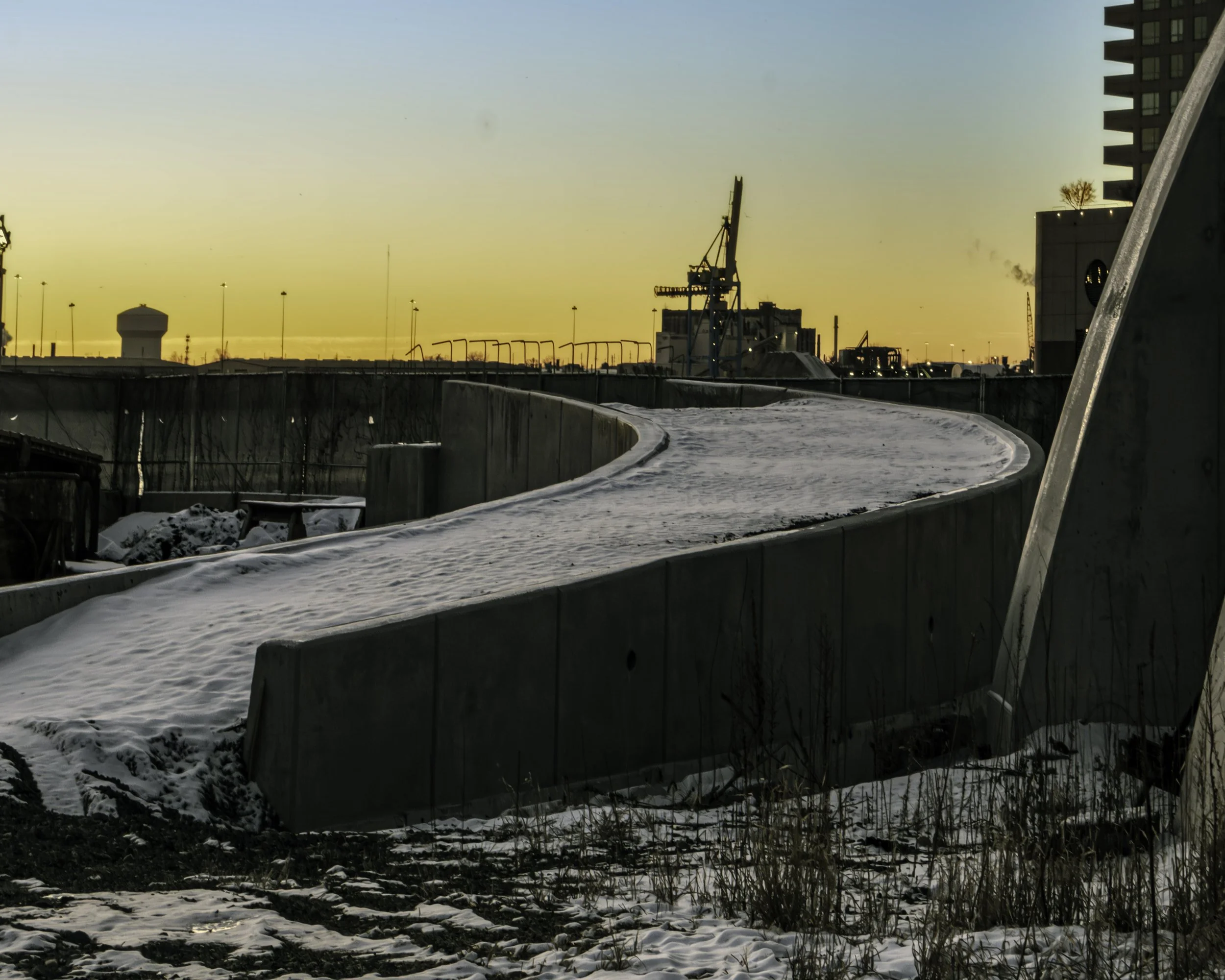 Cold Morning Bend
Snow-covered curved concrete path leads toward a waterfront industrial area under a pale winter sky.