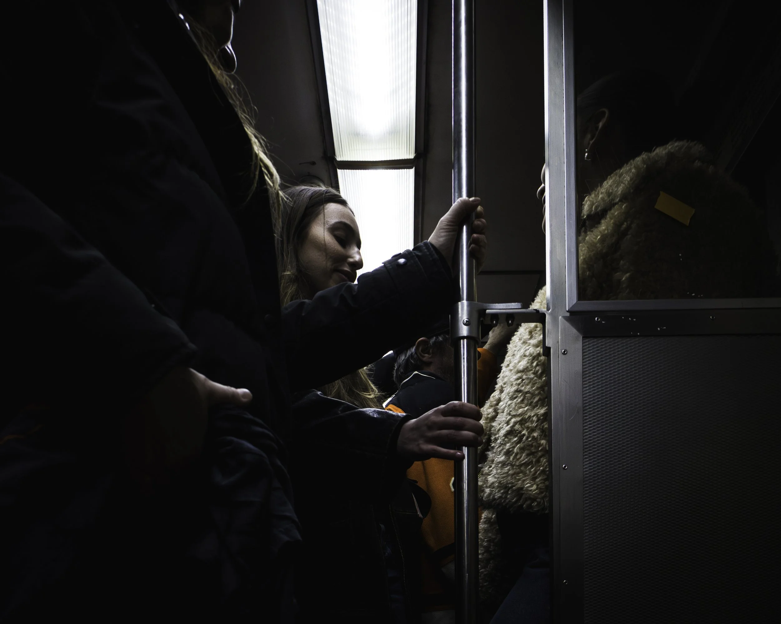 Transit Balance
Passenger grips a subway pole beneath a bright overhead light, surrounded by other riders in a dark train interior.