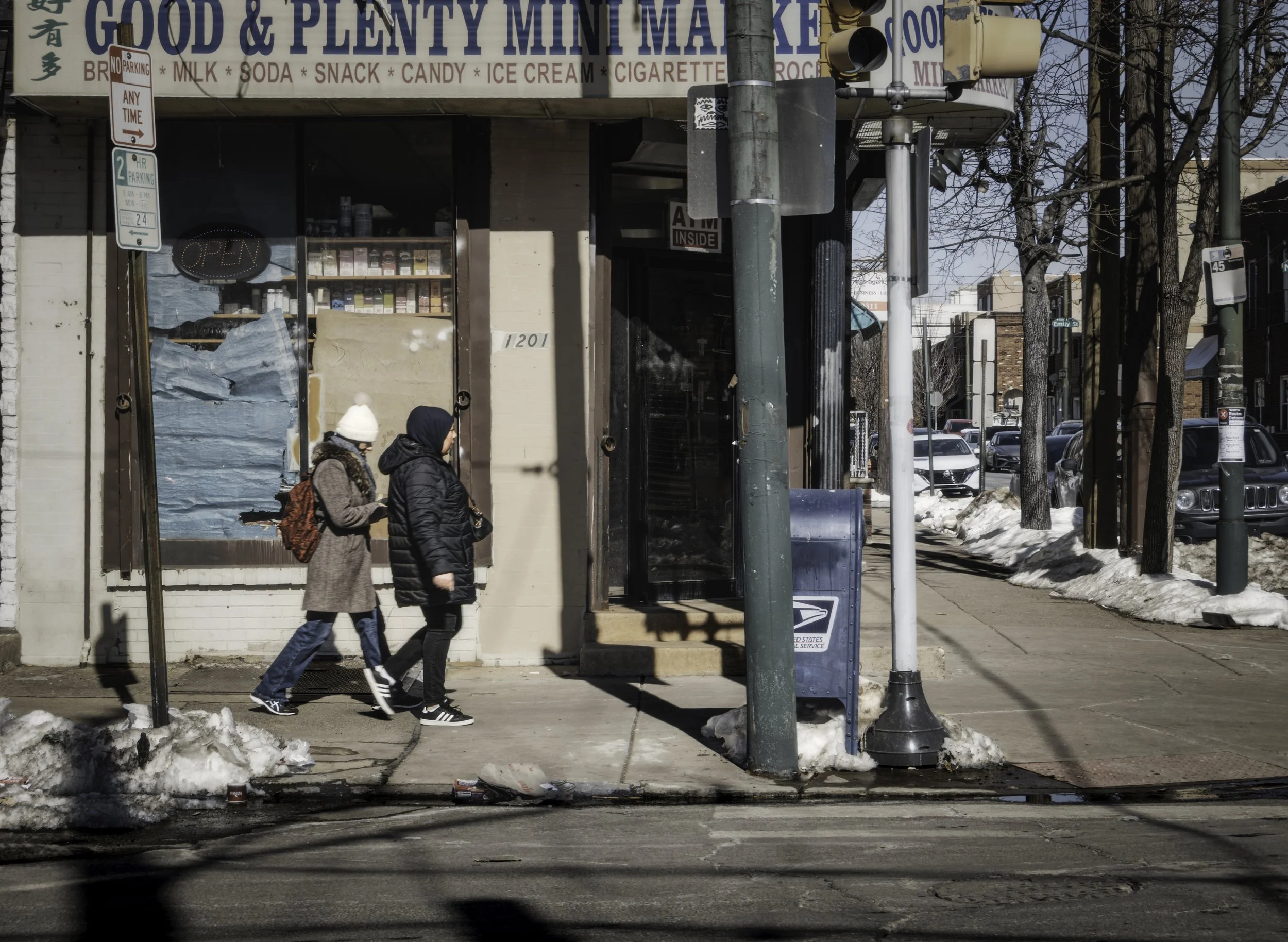 Snowbound Crossing
Two bundled pedestrians walk past a corner store with snow piled along the sidewalk and curb.