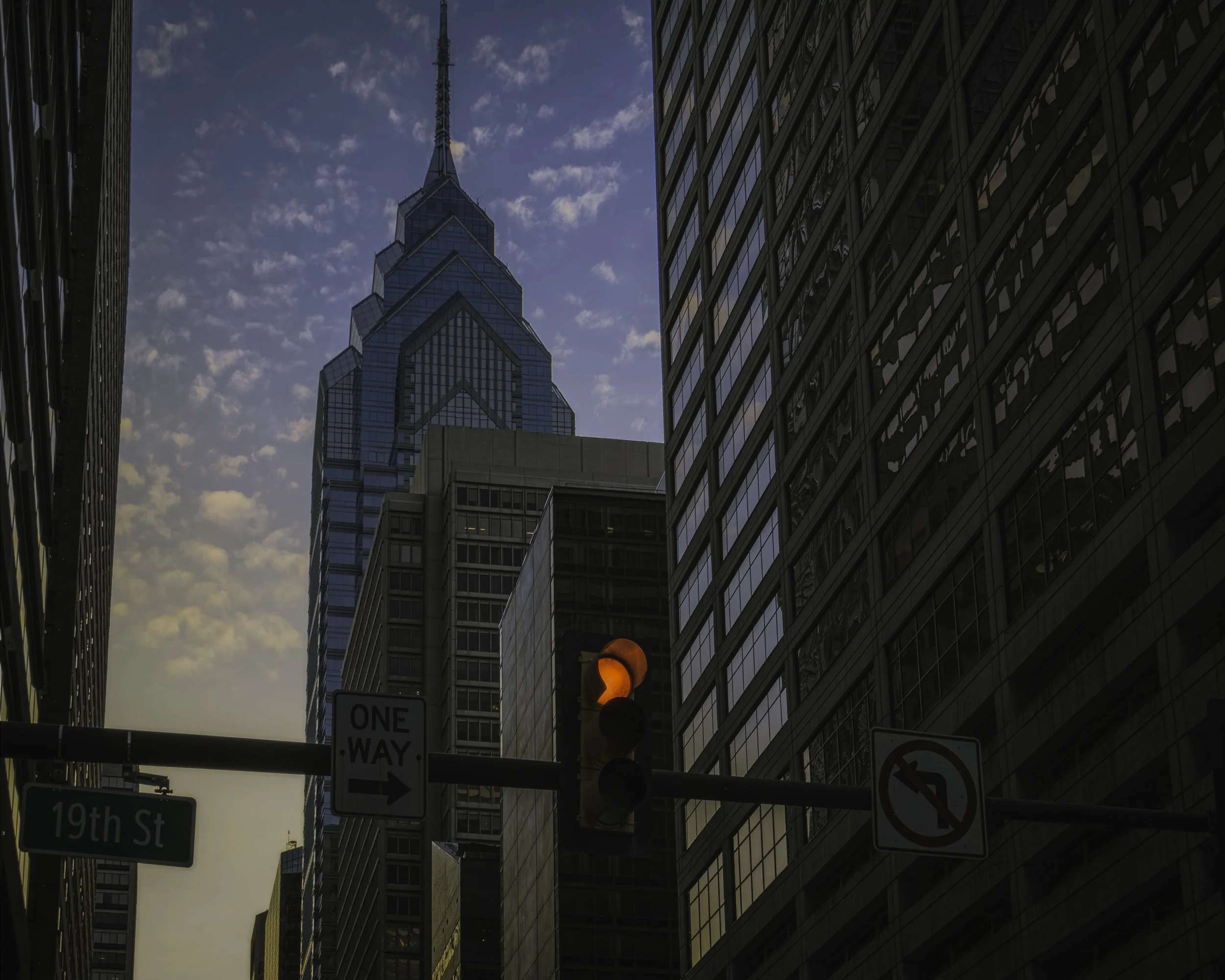 Tower and Signal
Skyscraper viewed from street level with a traffic light and city signs silhouetted against a cloudy sky.