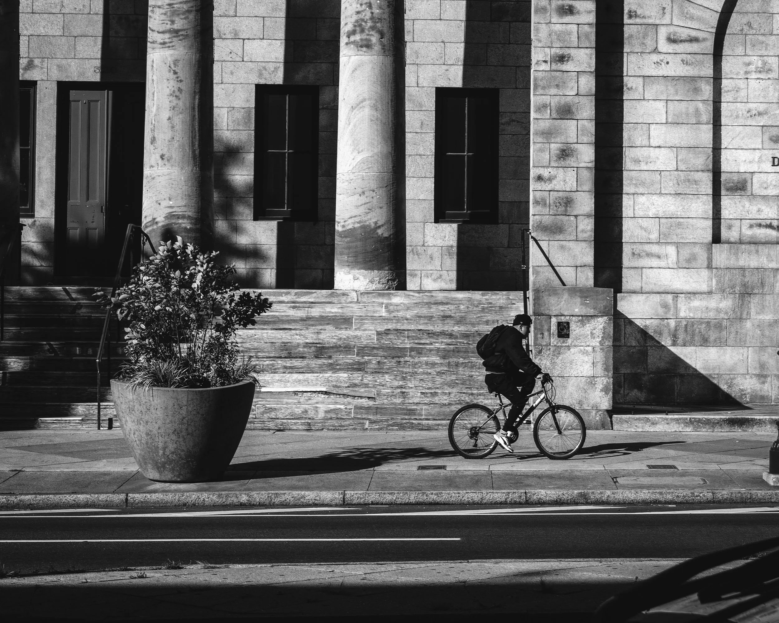 Lines of Light, Lines of Motion
Black and white street photograph on Philadelphia’s Avenue of the Arts, showing a cyclist passing a stone building with tall columns and a large planter.