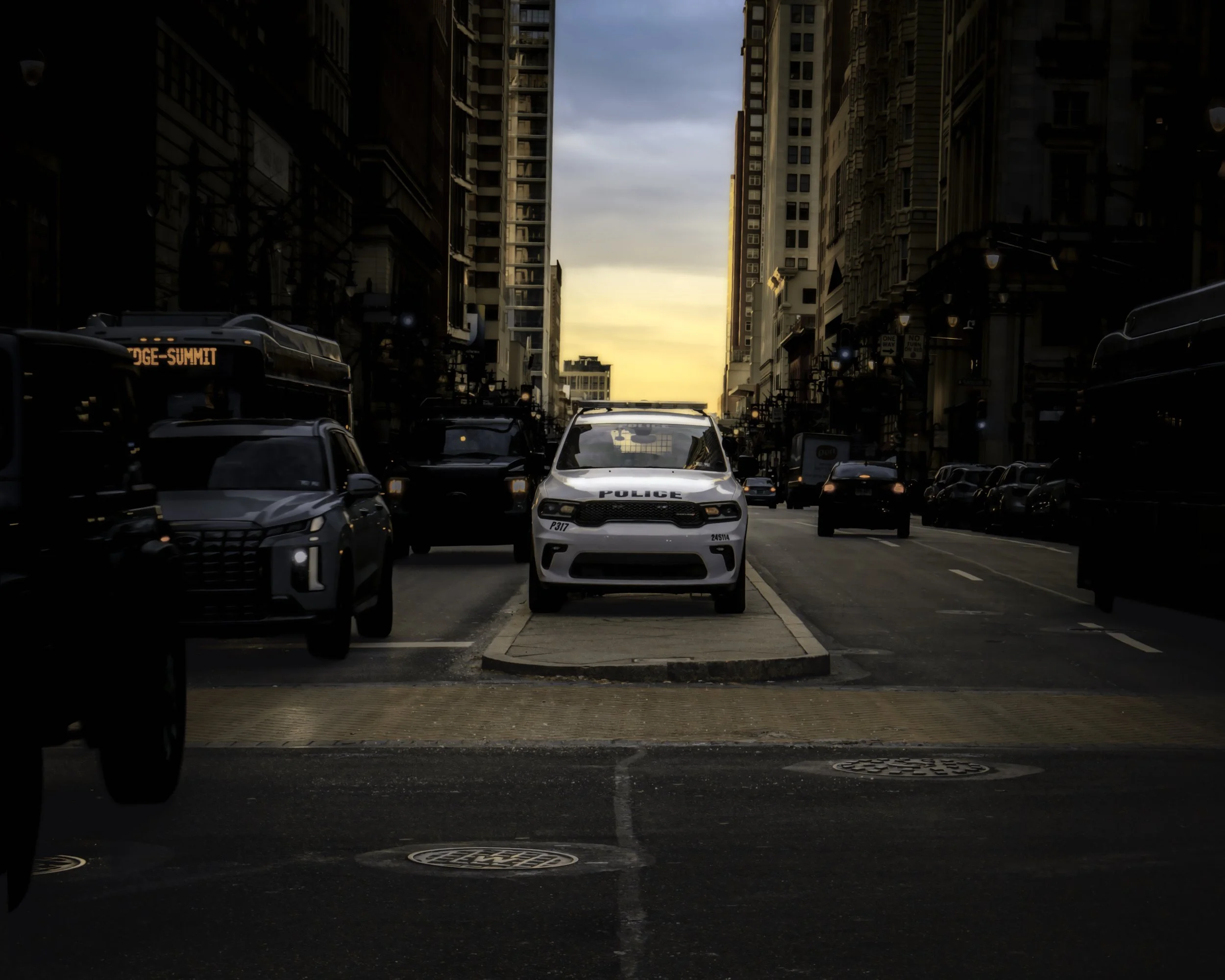 Median Patrol at Dawn
Philadelphia street at dawn with a white police SUV parked on a center median, framed by buses, traffic, and tall downtown buildings beneath a glowing morning sky.