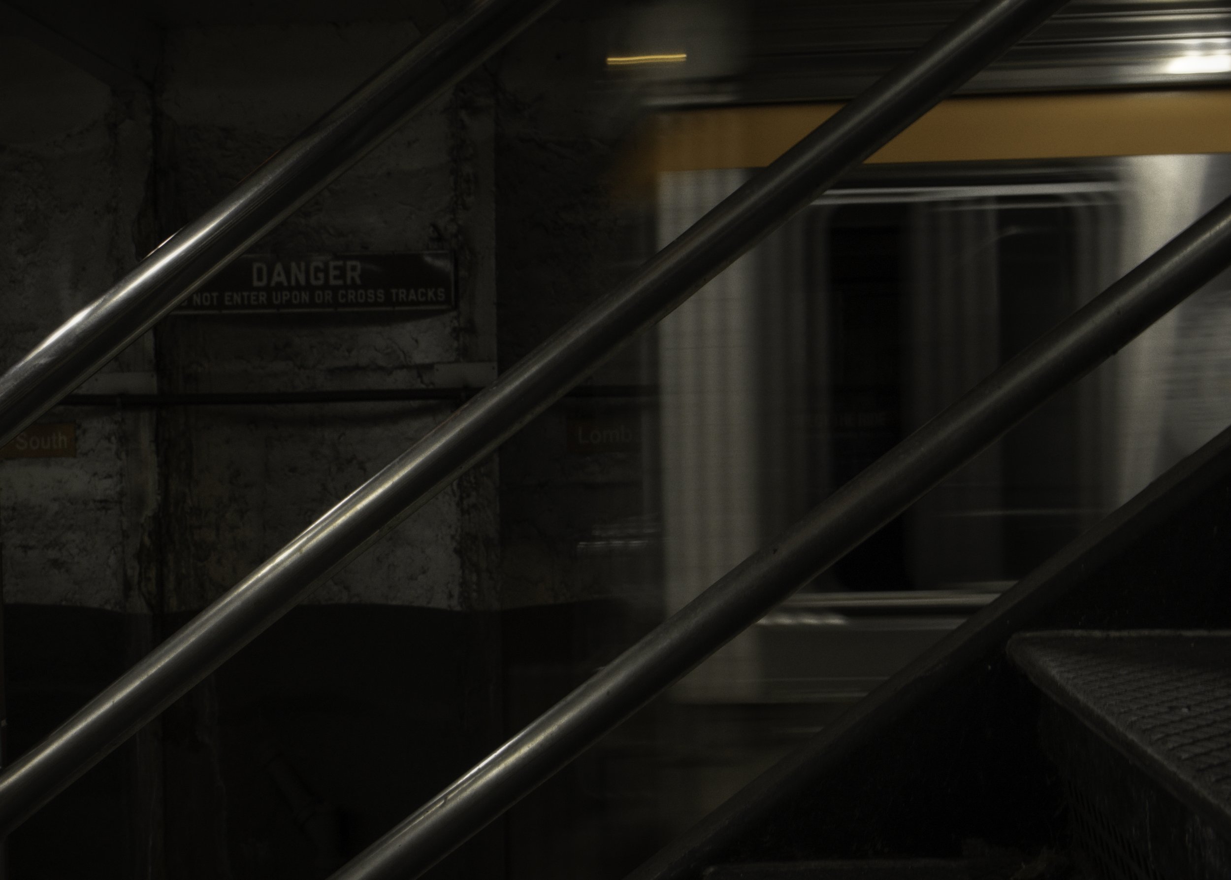 Diagonals and Motion
Blurred subway train passes behind diagonal metal railings, with a warning sign on the station wall in the background.