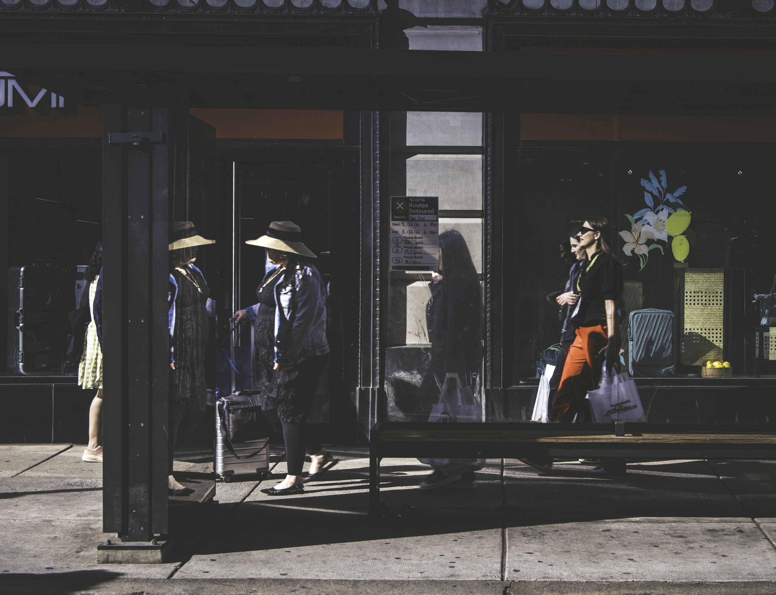 Three Versions of the Same Woman
A woman in a wide-brim hat and dark layered outfit enters a storefront, her figure multiplied across three glass door panels as reflection and reality blur together, while a second woman in orange trousers approaches 