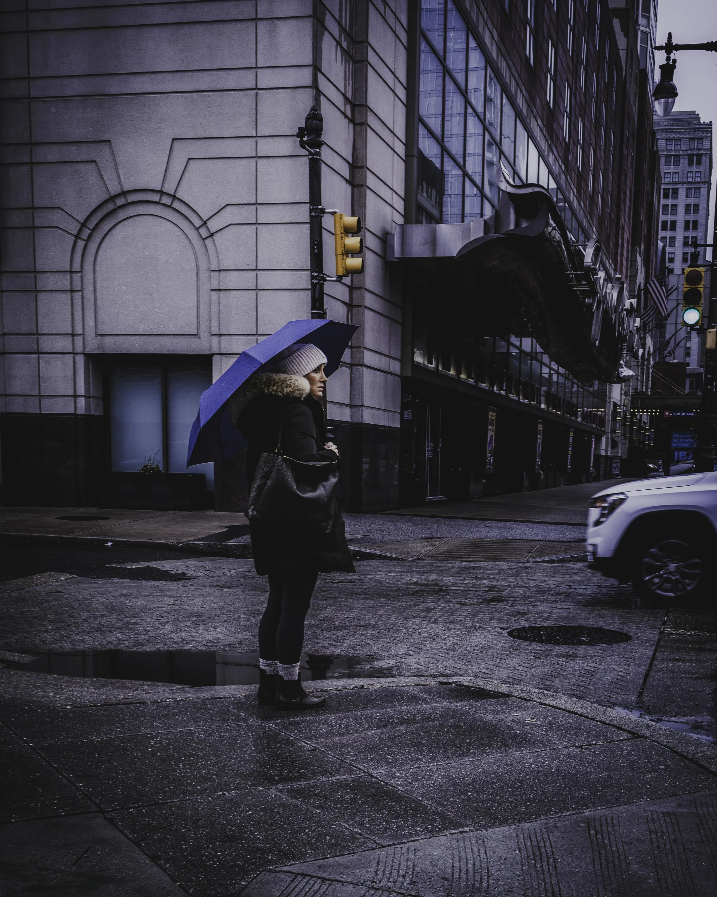 Waiting Under Blue
Woman waits at a wet street corner beneath a blue umbrella as a white vehicle enters the frame.
