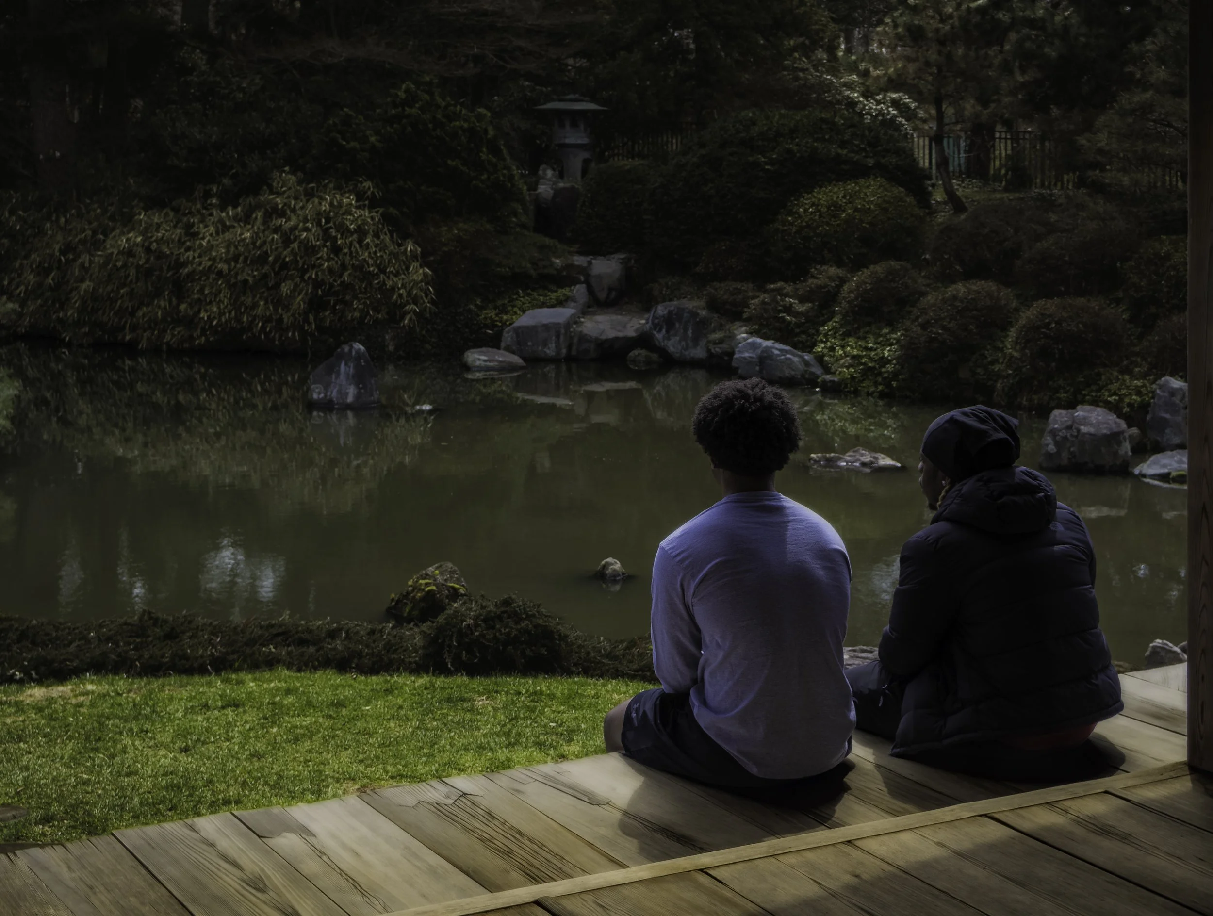 Quiet at the Garden
Two people sit on a wooden deck at Shofuso Japanese Cultural Center, overlooking a still pond surrounded by rocks, shrubs, and trees.