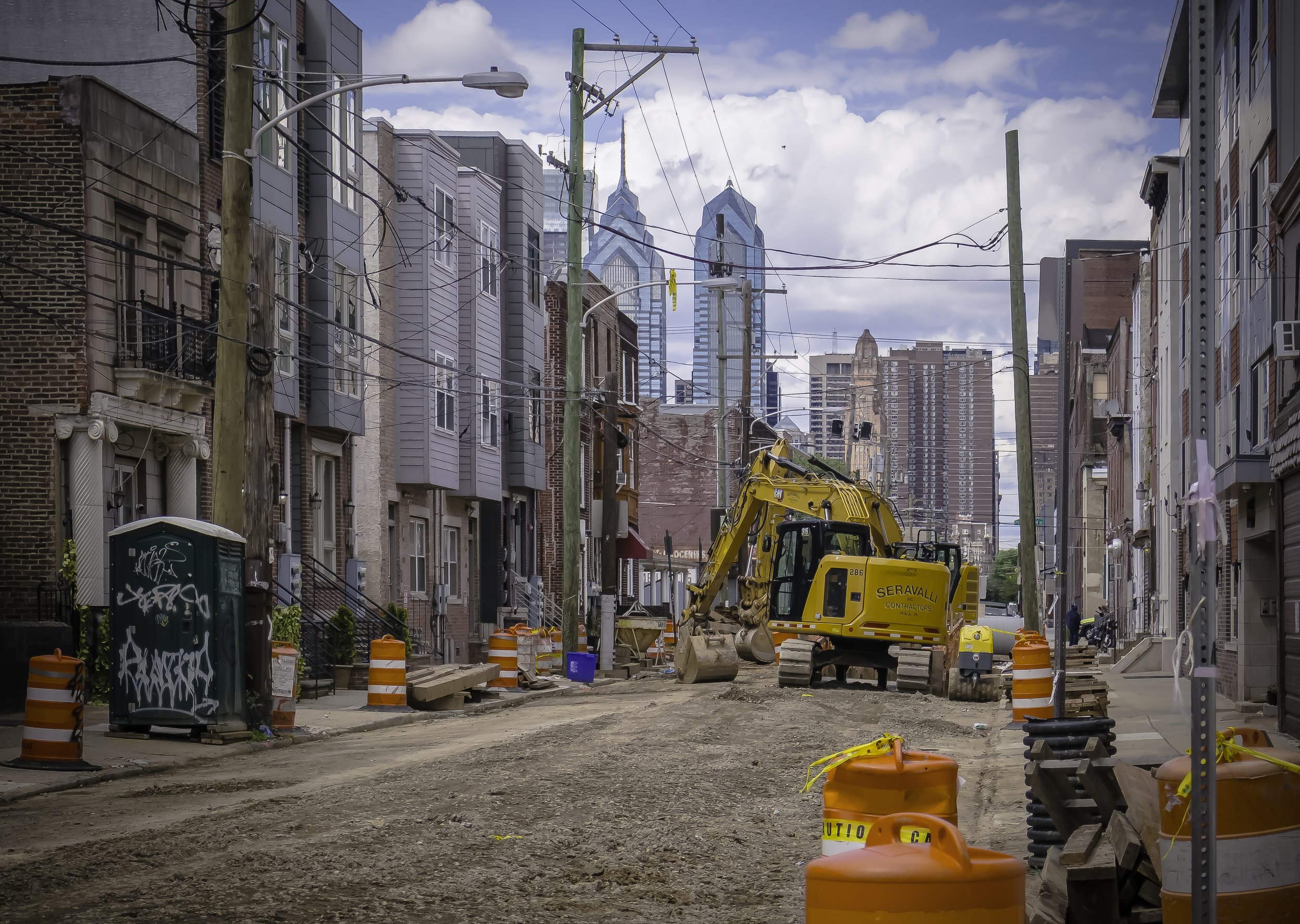 Orange Barrels, Blue Sky
Orange construction barrels and an excavator on a torn-up city street with the Philadelphia skyline in the distance.
