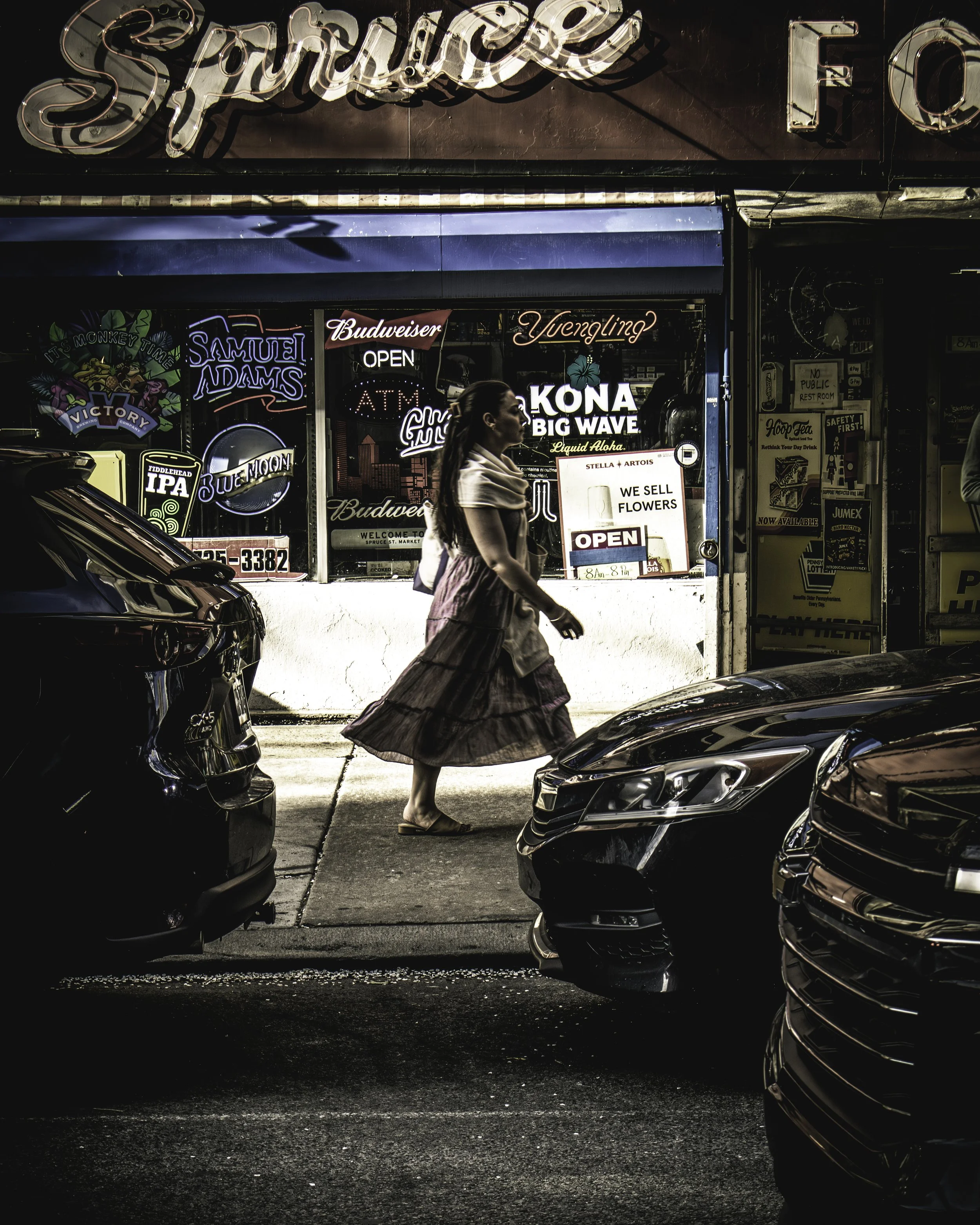 Spruce Street Passing
Woman in a flowing tiered skirt walks past a Philadelphia bar storefront on Spruce Street, framed between two dark parked cars, neon beer signs crowding the window behind her including Samuel Adams, Yuengling, and Kona Big Wave.
