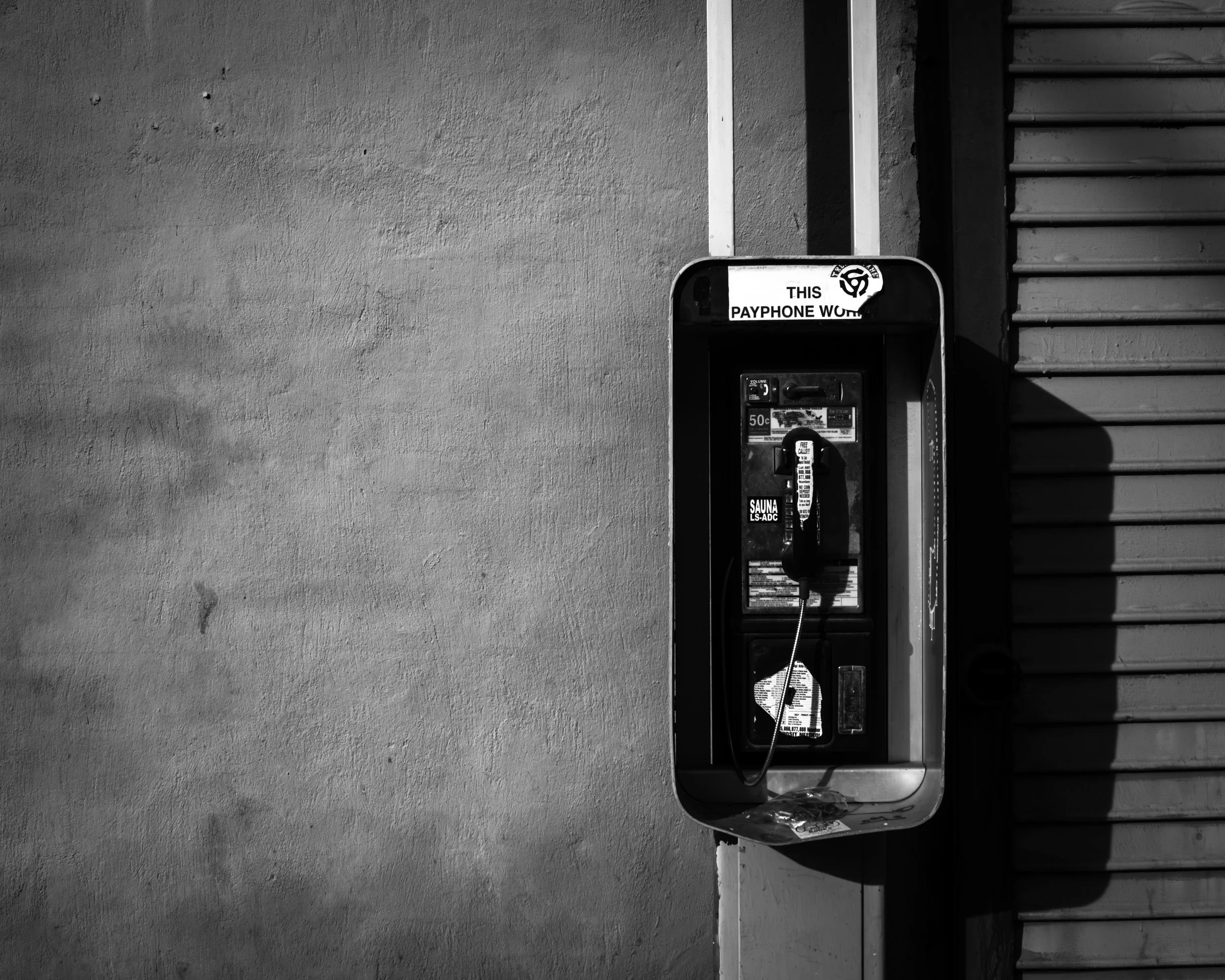 Echoes of a Lost Connection
Black and white payphone mounted on a plain wall beside a shutter, lit by strong sunlight and shadow.