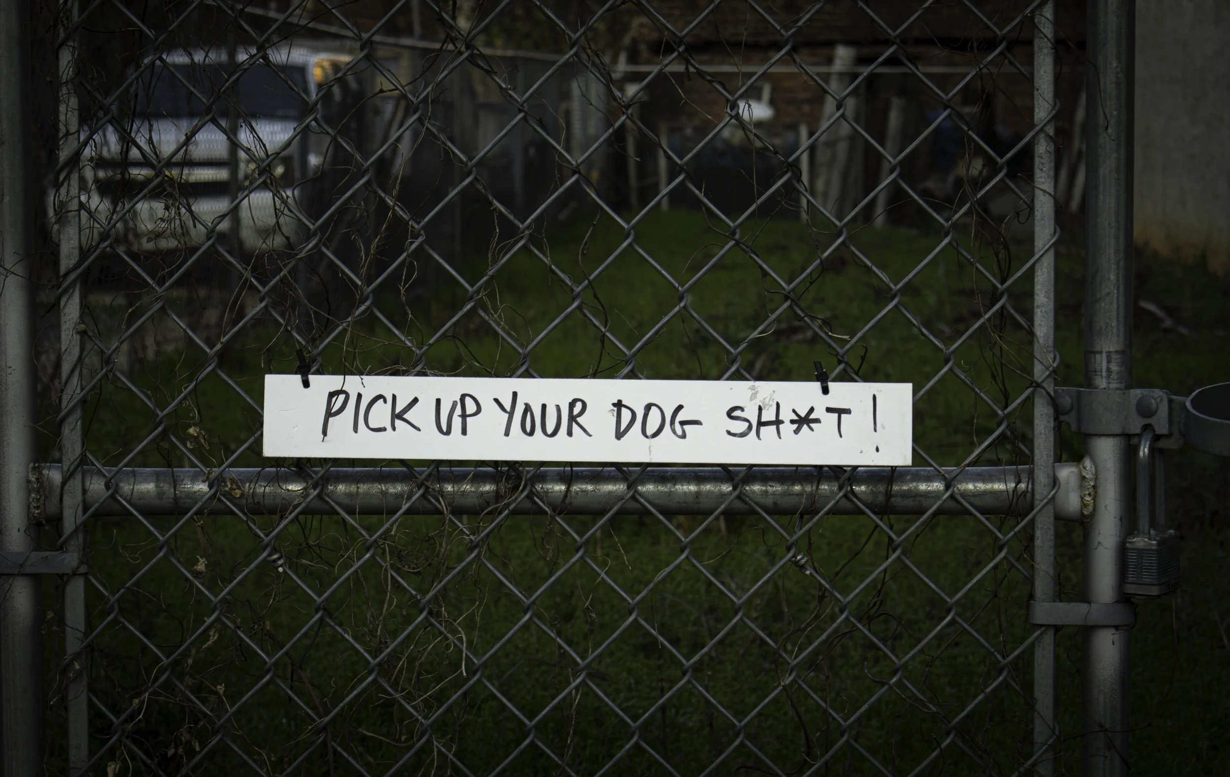Pick Up Your Dog Sh*t
Hand-lettered sign reading "PICK UP YOUR DOG SH*T!" zip-tied to a chain-link fence gate in a Philadelphia neighborhood, dark yard visible through the diamond mesh behind it. Philadelphia humor street photography, neighborhood si