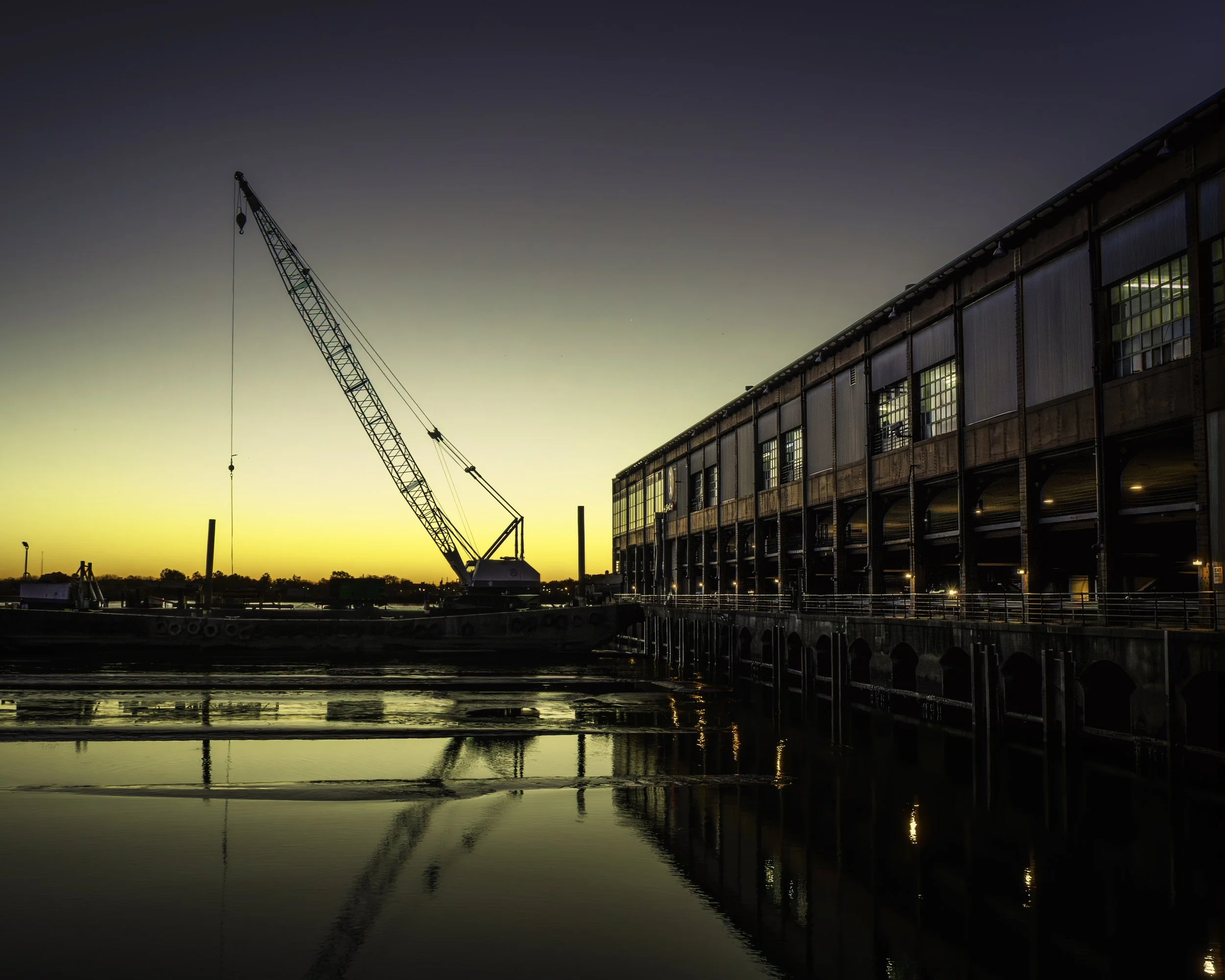 Echoes in the Slip
Crane and long pier building at dawn reflected in calm water, with window lights glowing along the waterfront structure.
