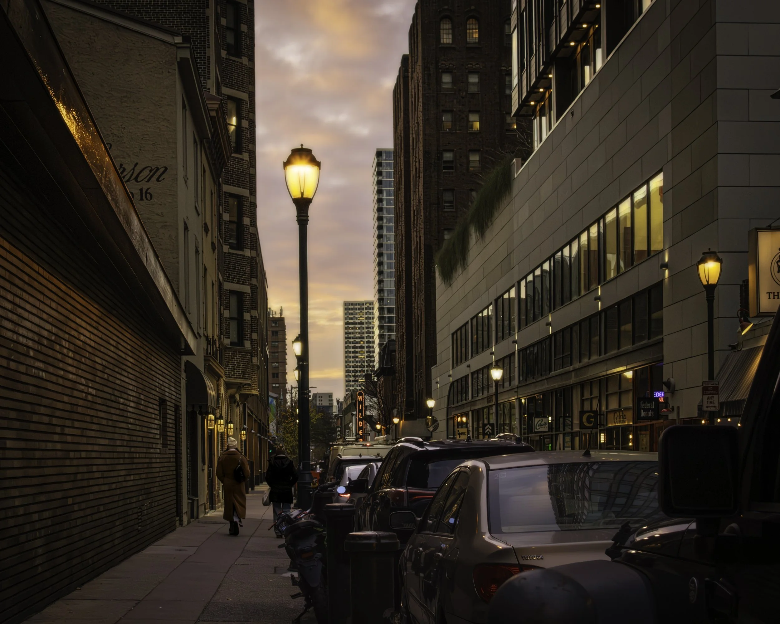 Last Walk of the Day
Narrow city sidewalk with parked cars, glowing streetlamps, and two pedestrians in evening light.