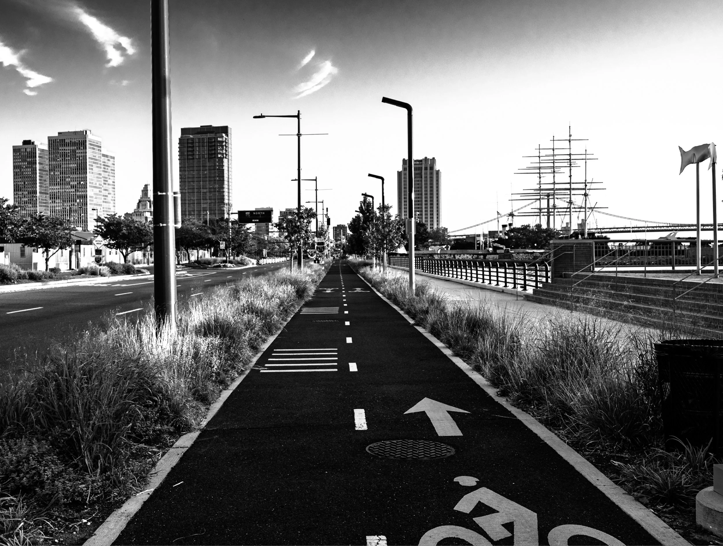 Black and white photograph of empty bike path leading toward city skyline with tall buildings, street lamps lining both sides, ship masts visible on right, urban landscape with dramatic sky