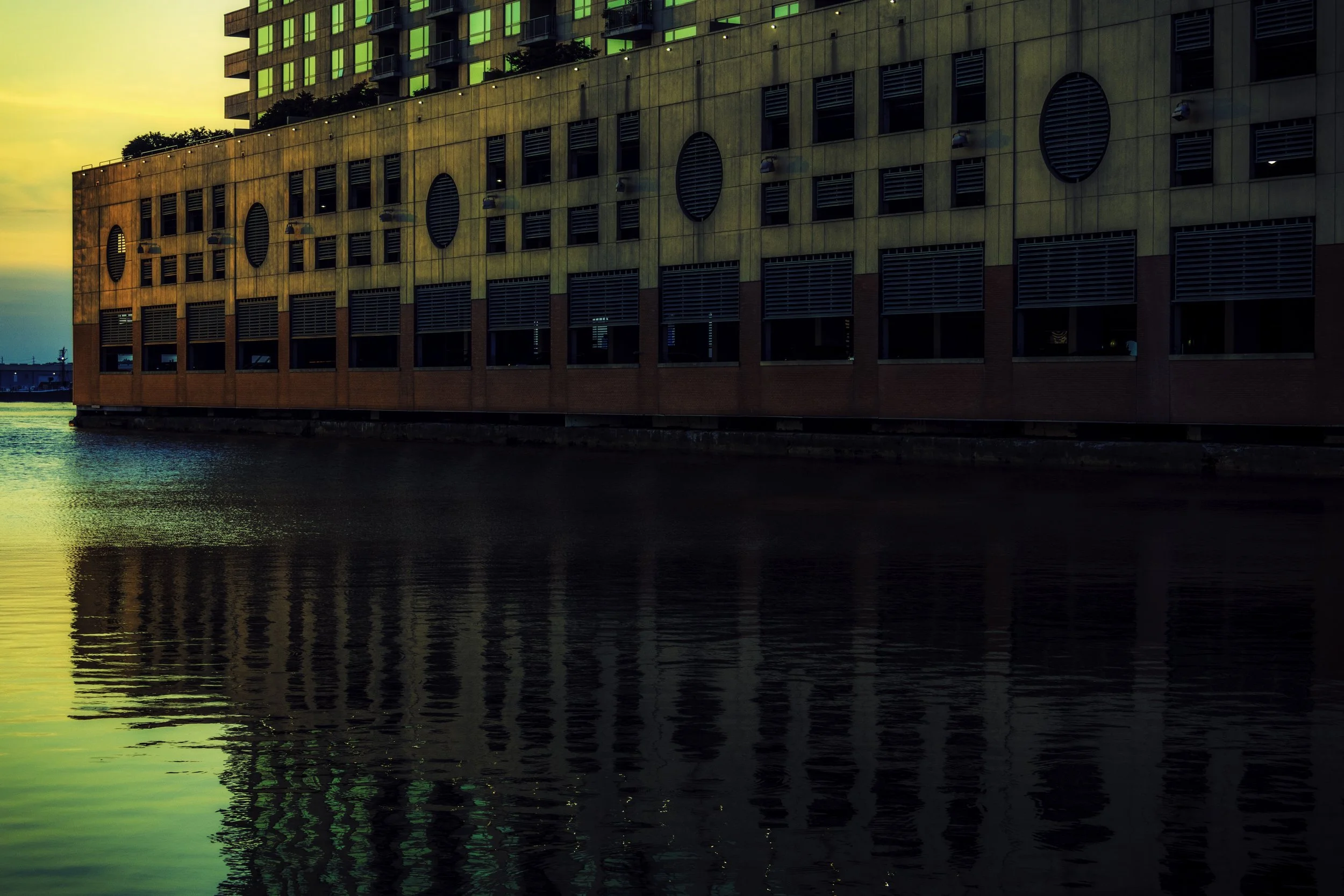 Reflections Along the Wall
Waterfront building with circular window openings reflected in dark water at dusk, with apartment windows glowing above the façade.