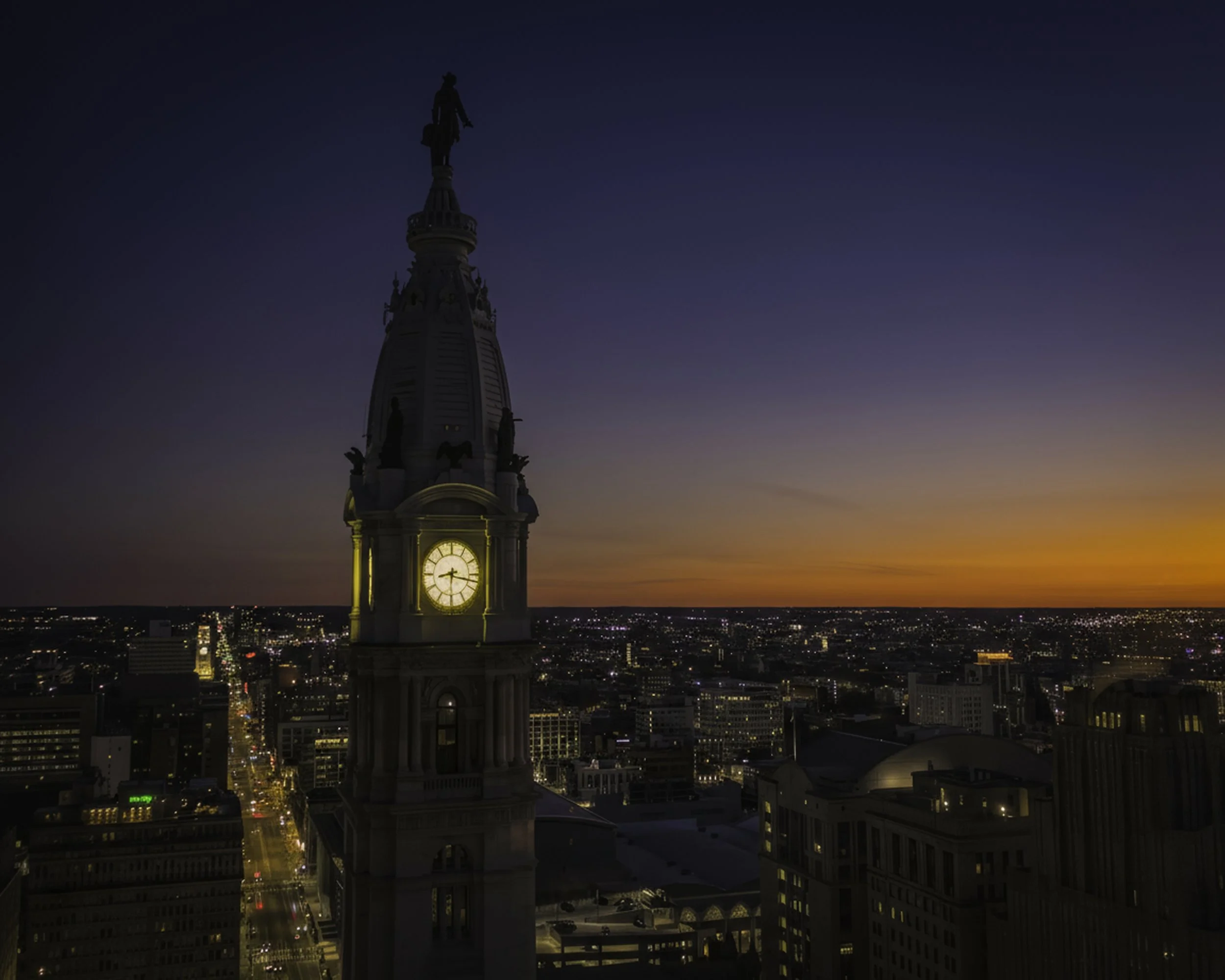 City Hall at First Light
A twilight aerial view of Philadelphia City Hall clock tower with the city grid glowing below under a fading sunset horizon.
