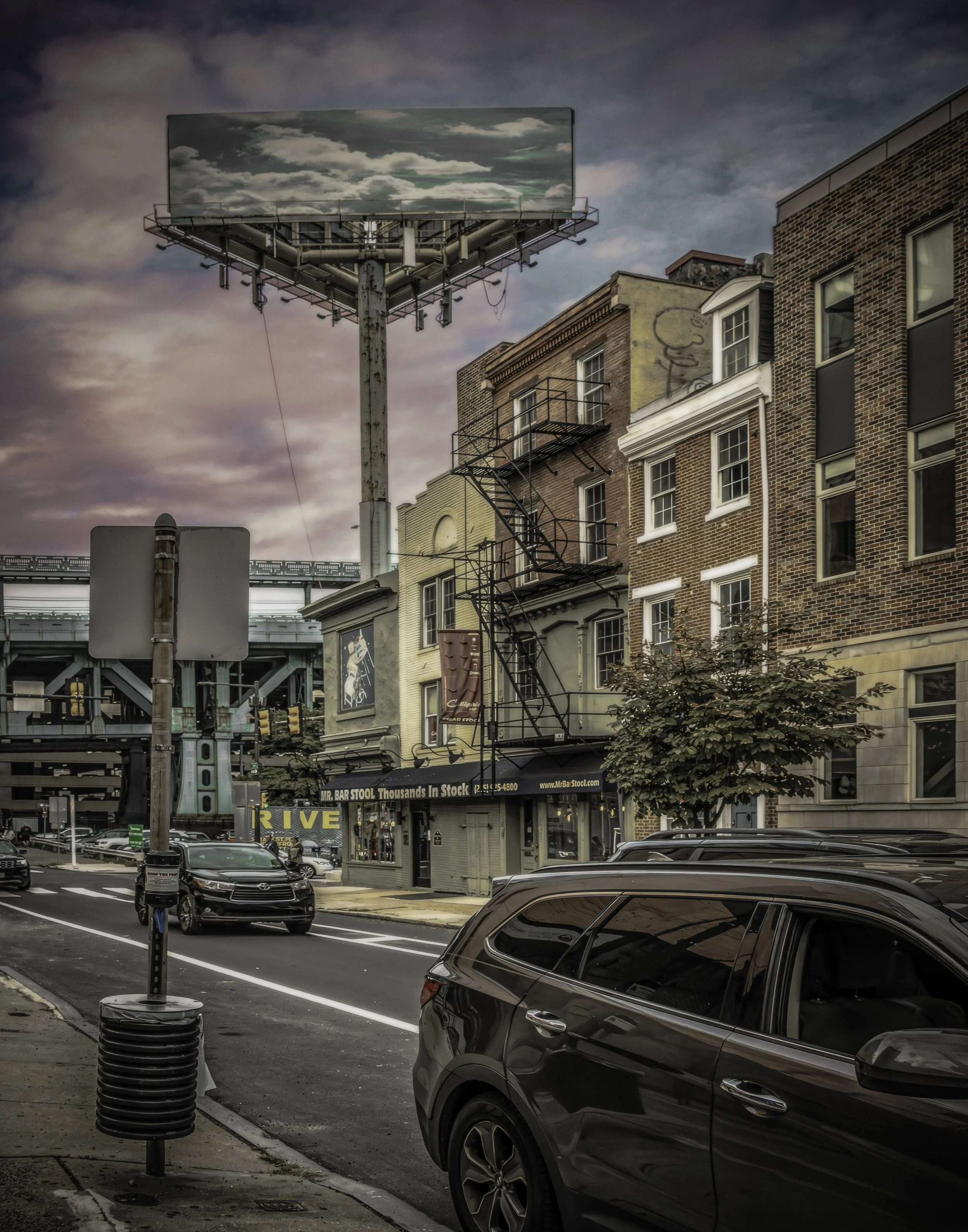 Billboard Over the Block
Billboard above 2nd Street in Philadelphia, rising over brick buildings, parked cars, and storefronts, with the Benjamin Franklin Bridge structure visible in the background.