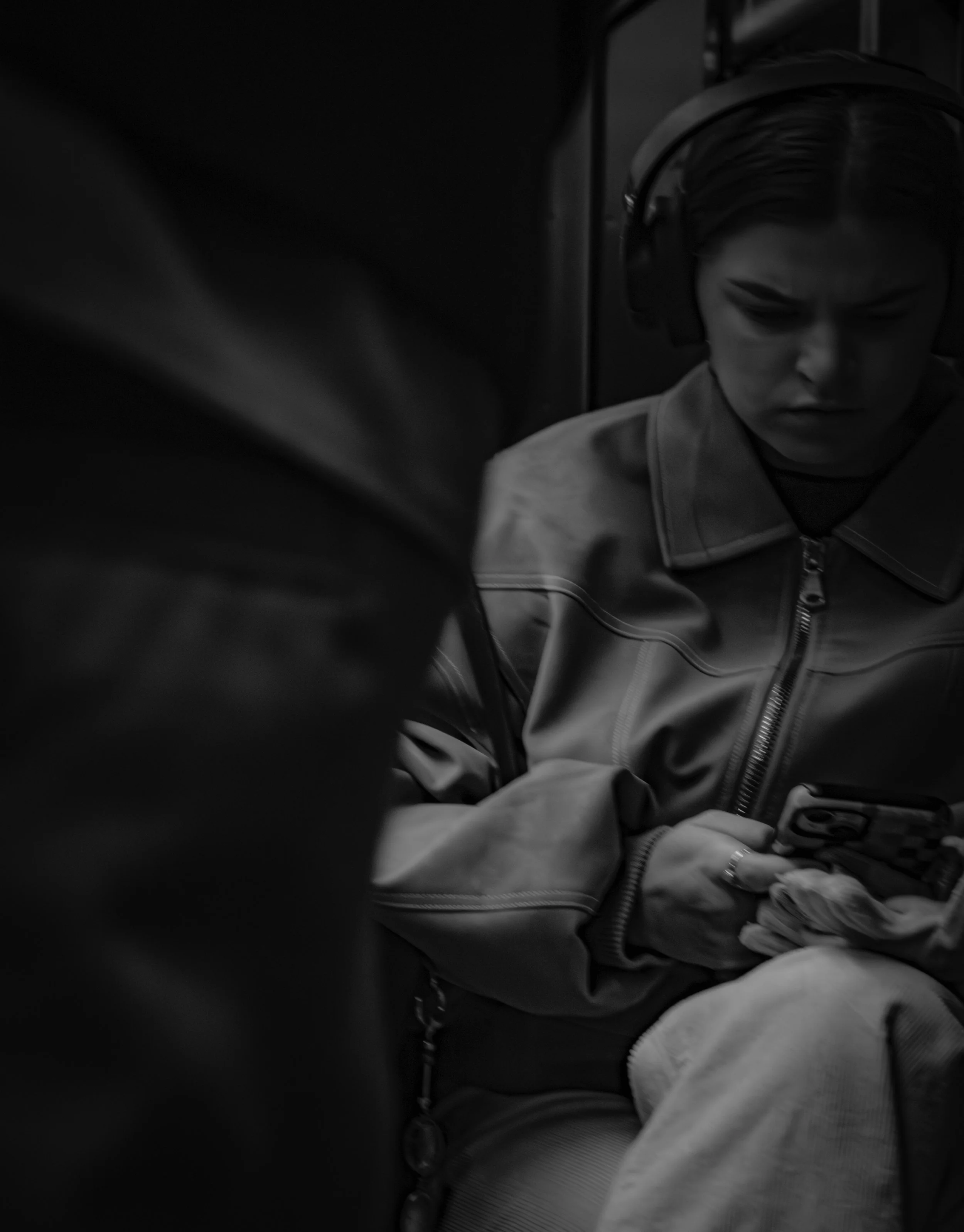 Inside the Quiet
Black-and-white close-up of a young woman wearing headphones on public transit, looking down at her phone with a serious, inward expression.