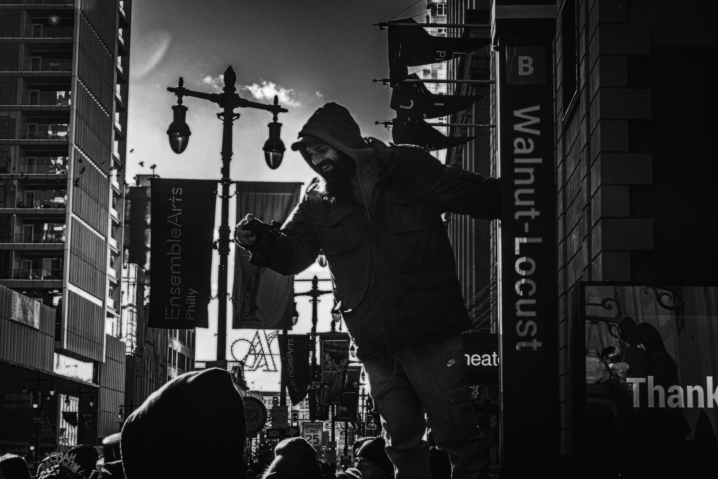 Leaning Into Light
Man holding a camera leans forward above a crowd on a sunlit downtown street on New Year’s Day, with banners and signs in the background.