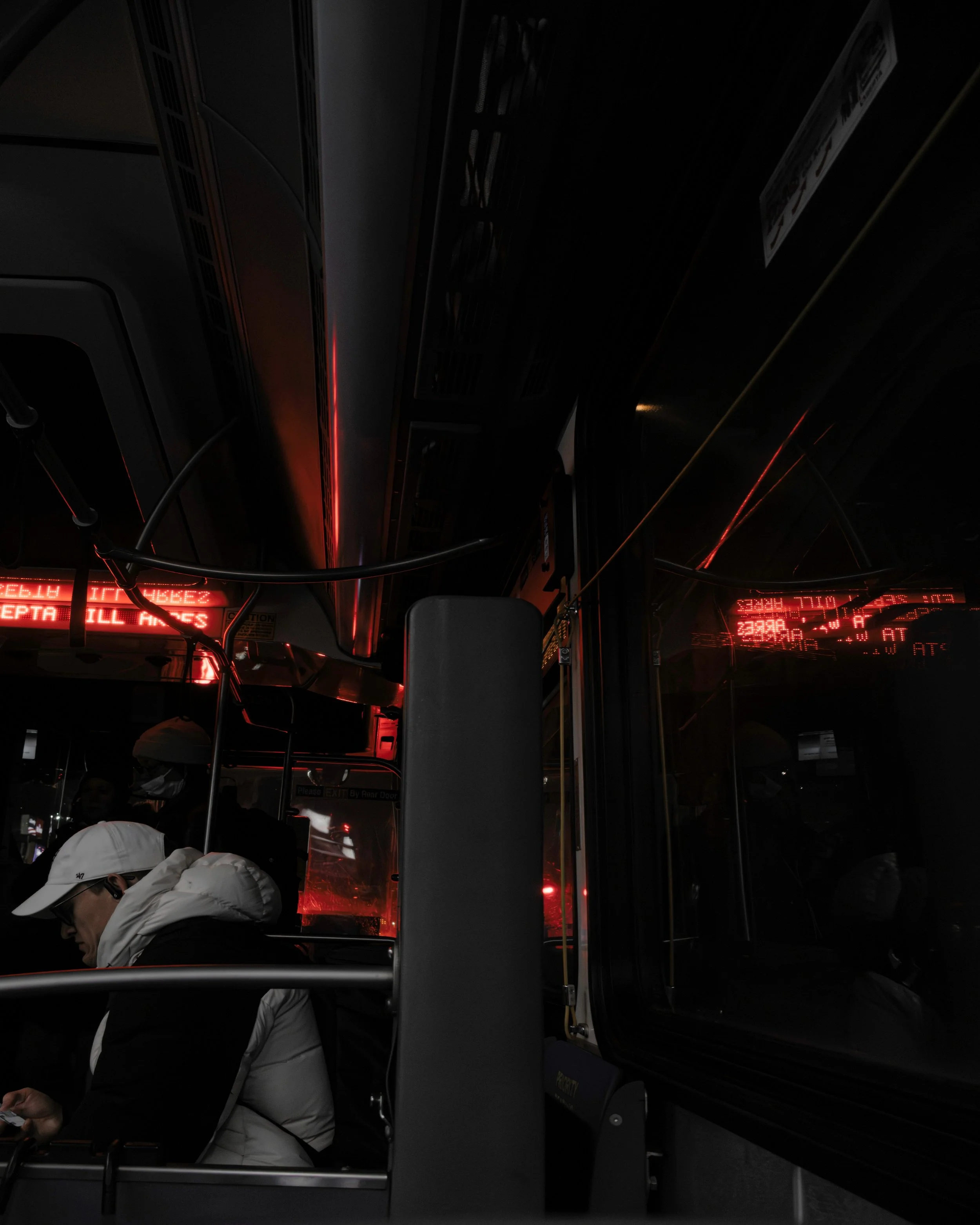 Reflections on the Night Bus
Dark city bus interior with a seated passenger in a white jacket, red LED route lights glowing overhead and reflected in the window.
