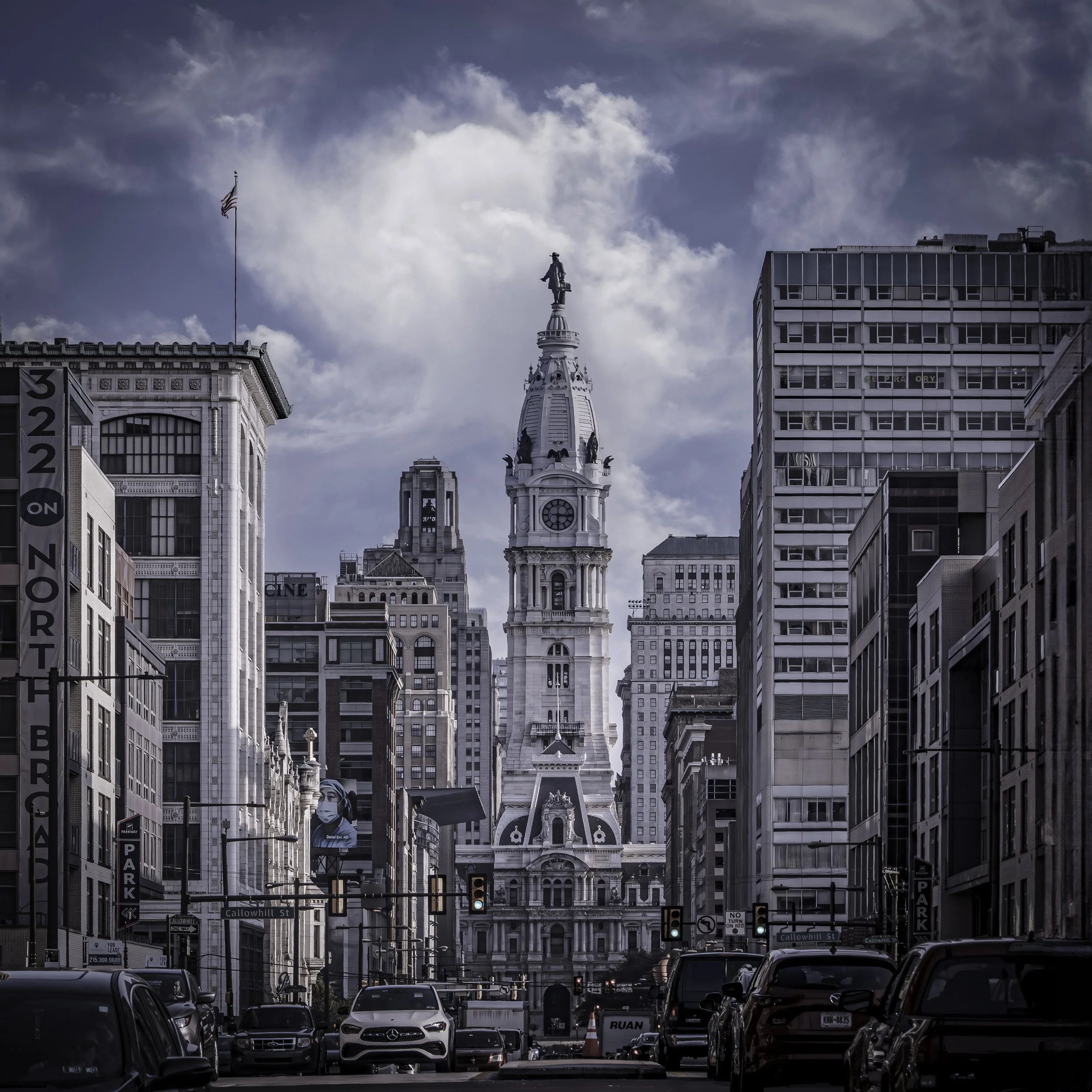 Penn at the End of Broad
Philadelphia City Hall tower rises at the vanishing point of North Broad Street, William Penn's statue silhouetted at its peak against a dramatic sky of billowing white clouds, the historic and modern buildings of the city ca
