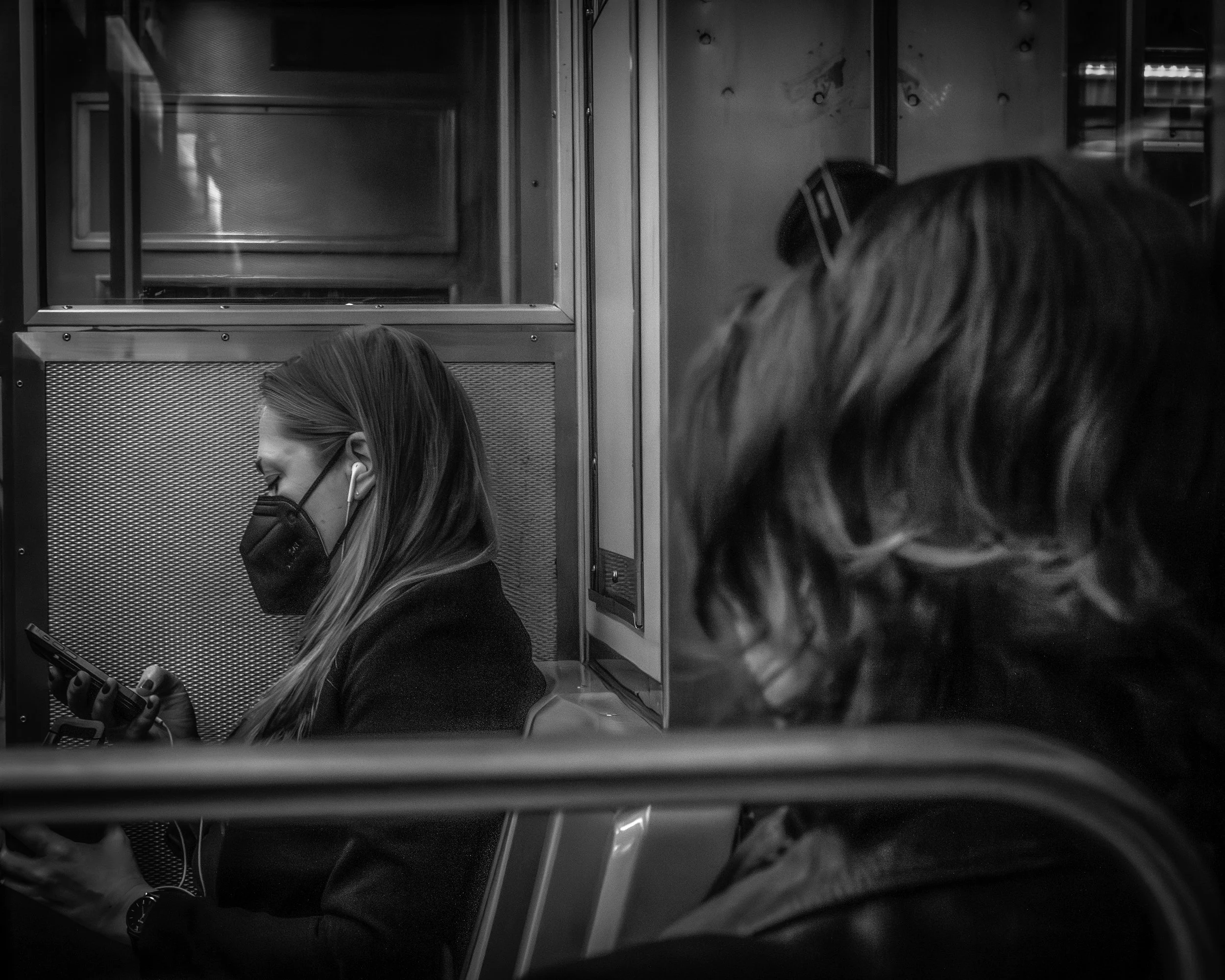 Moment in Transit
Black and white subway interior with a masked passenger looking at a phone while another rider is blurred in the foreground.