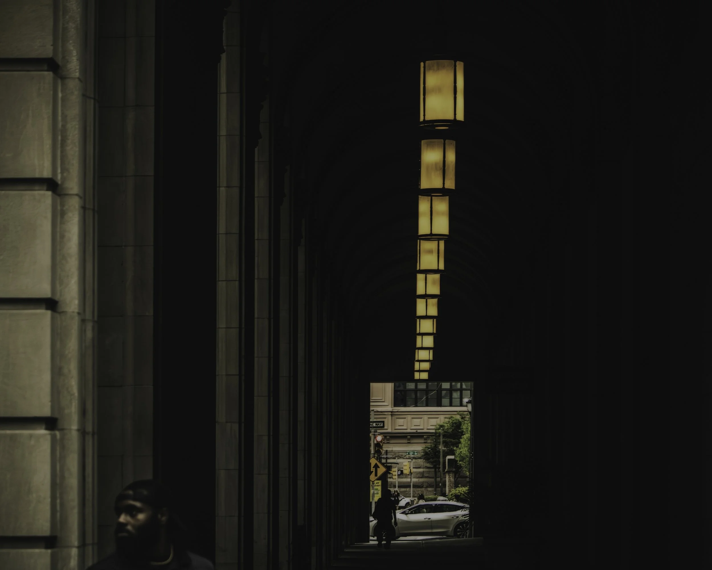 The Liminal Corridor
Dark architectural corridor with repeating overhead lights framing a distant street view and a shadowed figure in the foreground.