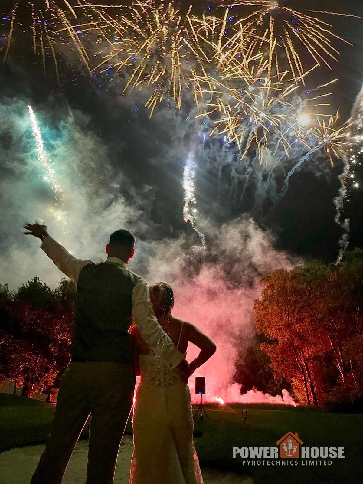 A couple watching fireworks display at night, with the man pointing towards the fireworks and the woman standing beside him. Smoke and colorful explosions light up the sky, and there is a logo for Power House Pyrotechnics Limited in the bottom right 