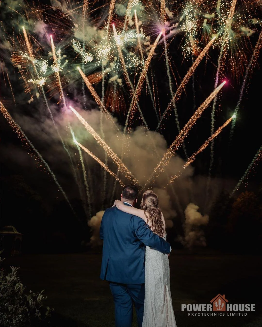 A couple in formal attire watching a fireworks display at night, with fireworks lighting up the sky.