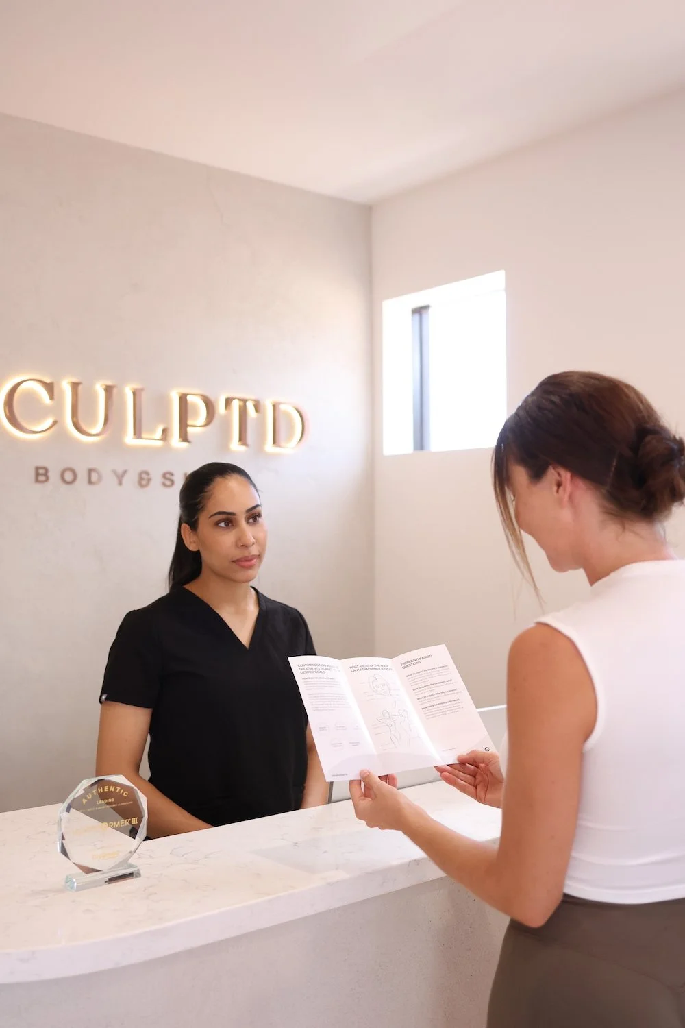 A woman at a reception desk holding informational pamphlet, speaking to a receptionist at CULPTD Body & Skin Center, with illuminated sign behind them reading 'CULPTD'.