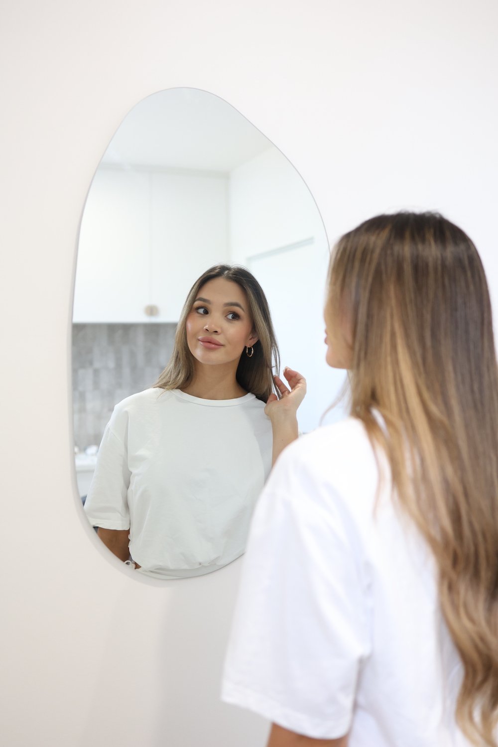 A woman with light brown hair looking at herself in a mirror on a white wall, touching her hair, in a bright room.