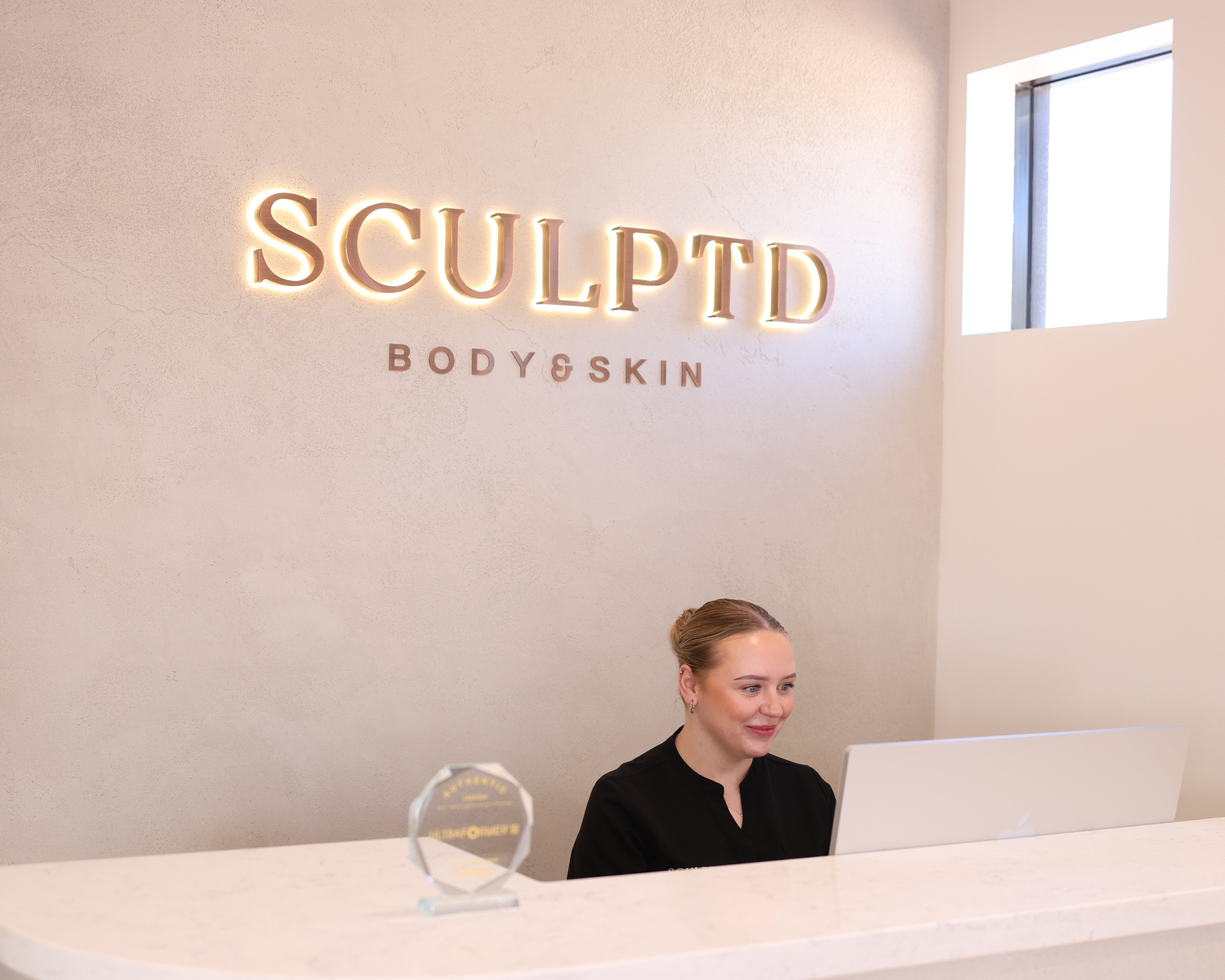 A woman sitting at a reception desk with a computer in front of her, inside a business called 'SCULPTD BODY & SKIN'. There is a sign with the business name illuminated on the wall behind her, and a small award plaque on the desk.